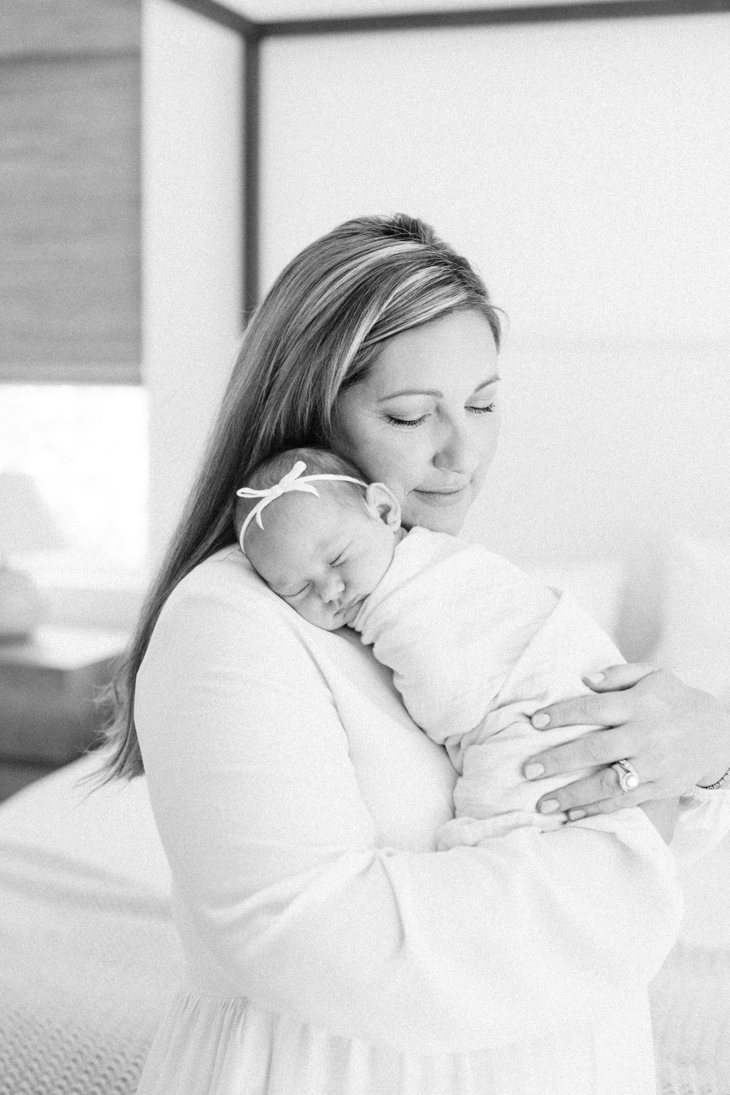 Black and white photo of a mother in white dress tenderly holding her swaddled newborn baby girl with a bow.