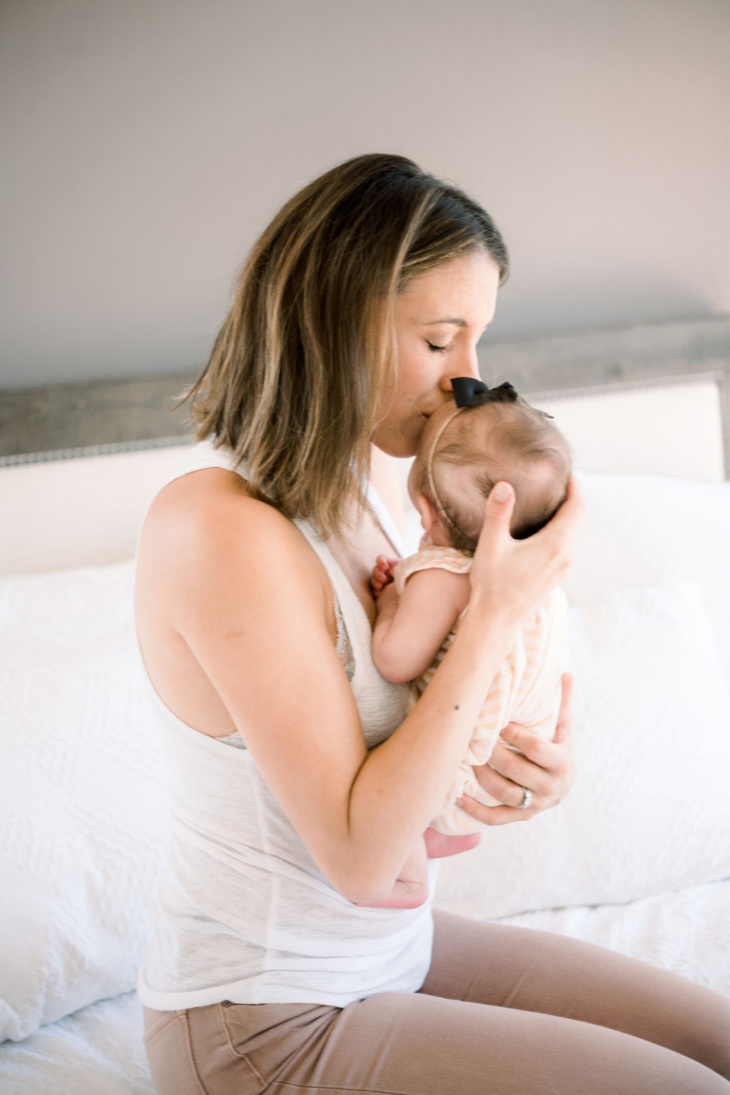 Loving mother kissing her newborn baby girl with black bow while sitting on white bed.