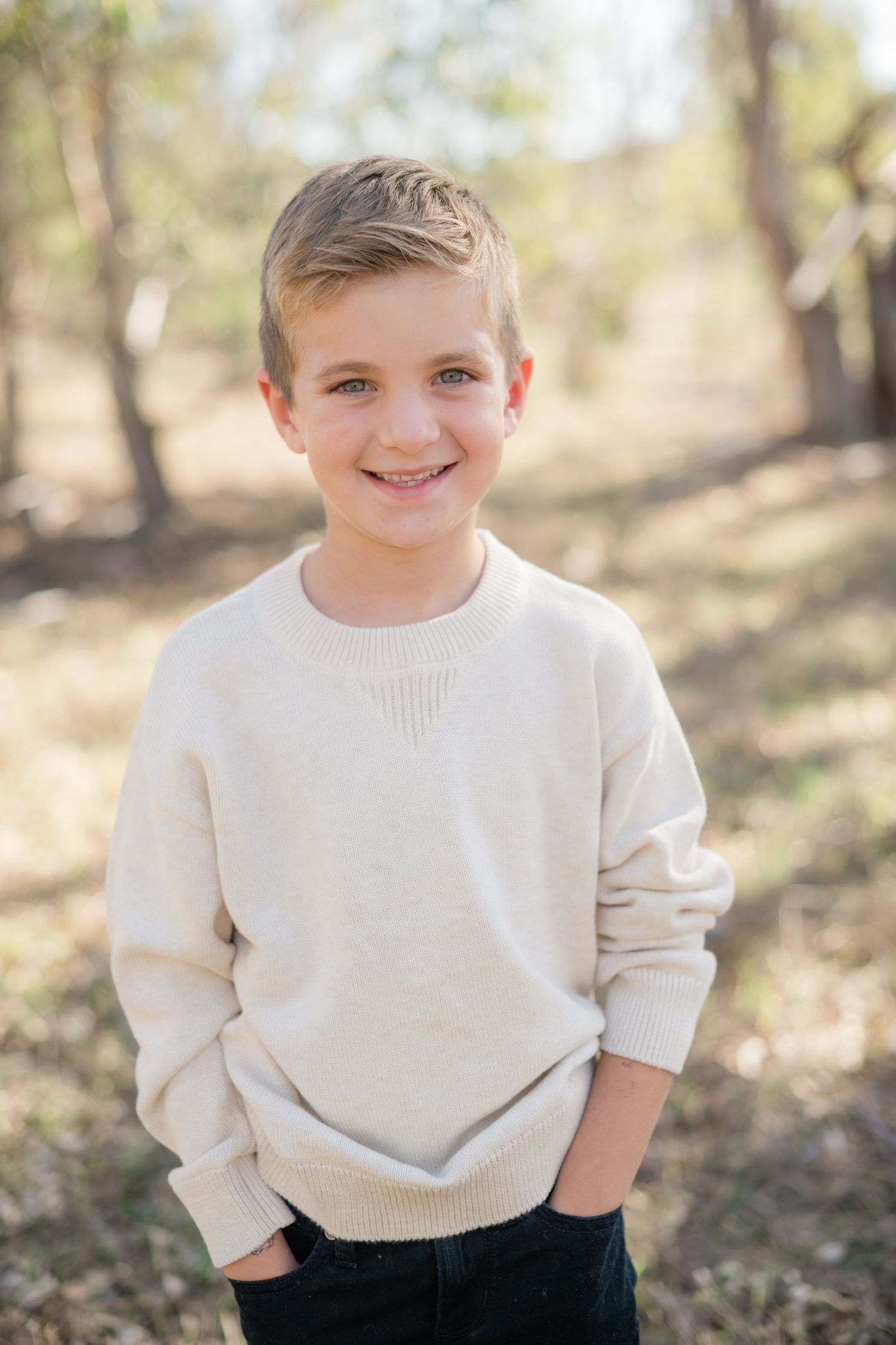 Smiling boy in a cream sweater with hands in pockets, standing outdoors in a sunlit wooded area.