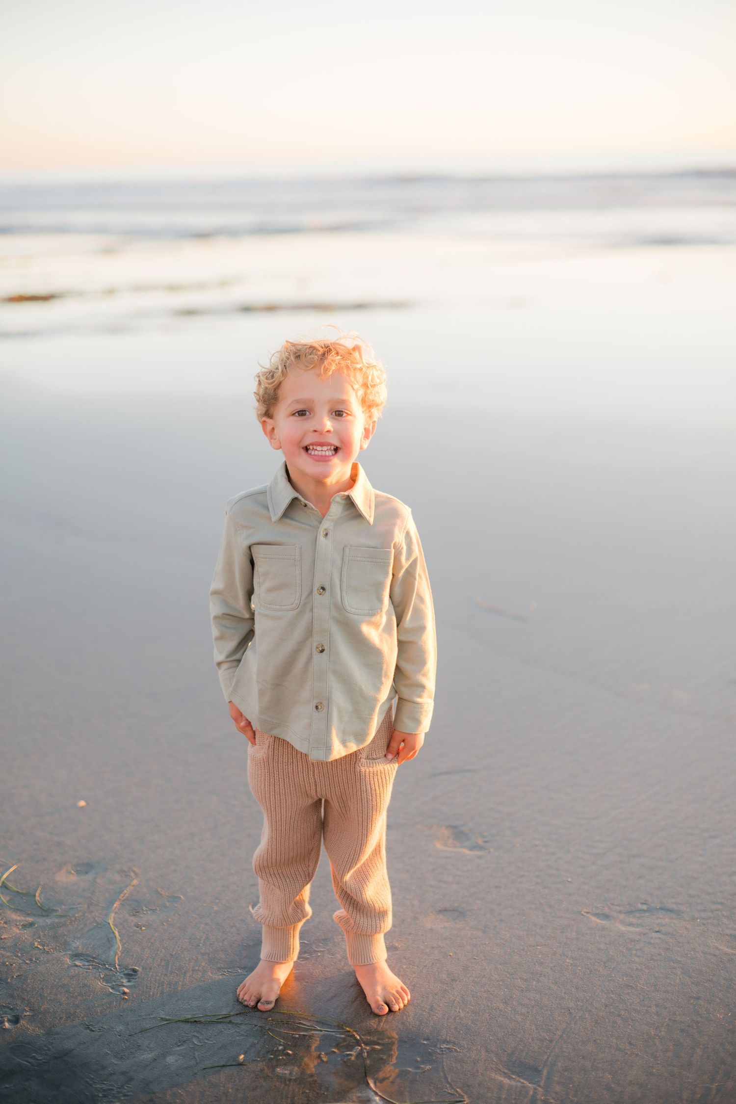 A smiling young boy with curly hair stands barefoot on a sandy beach at sunset wearing a sage shirt and tan pants.
