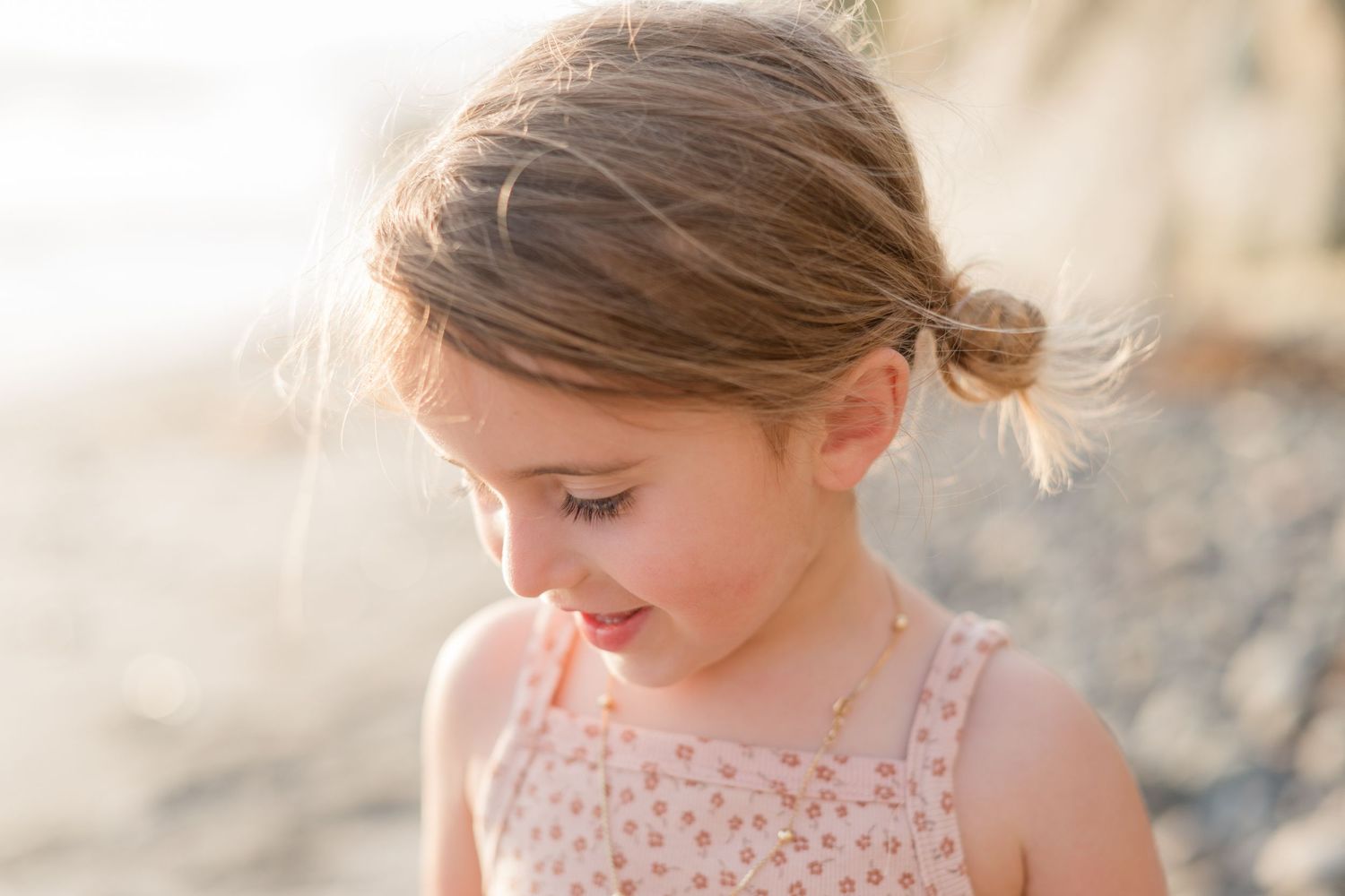 Young girl with hair bun in pink dress looks down at beach in golden sunlight.