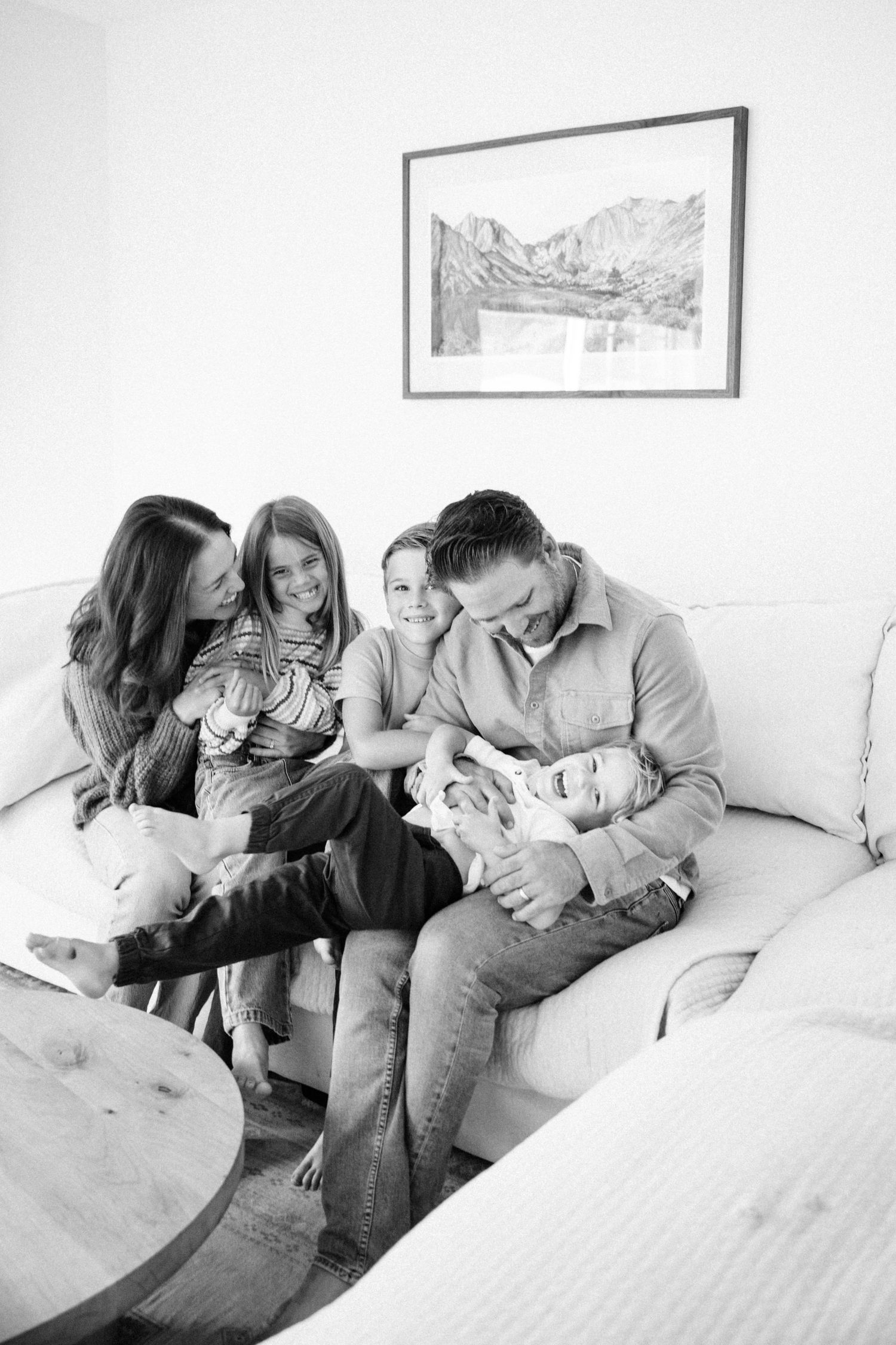 Black and white photo of a joyful family of five cuddling and laughing together on a white couch at home.