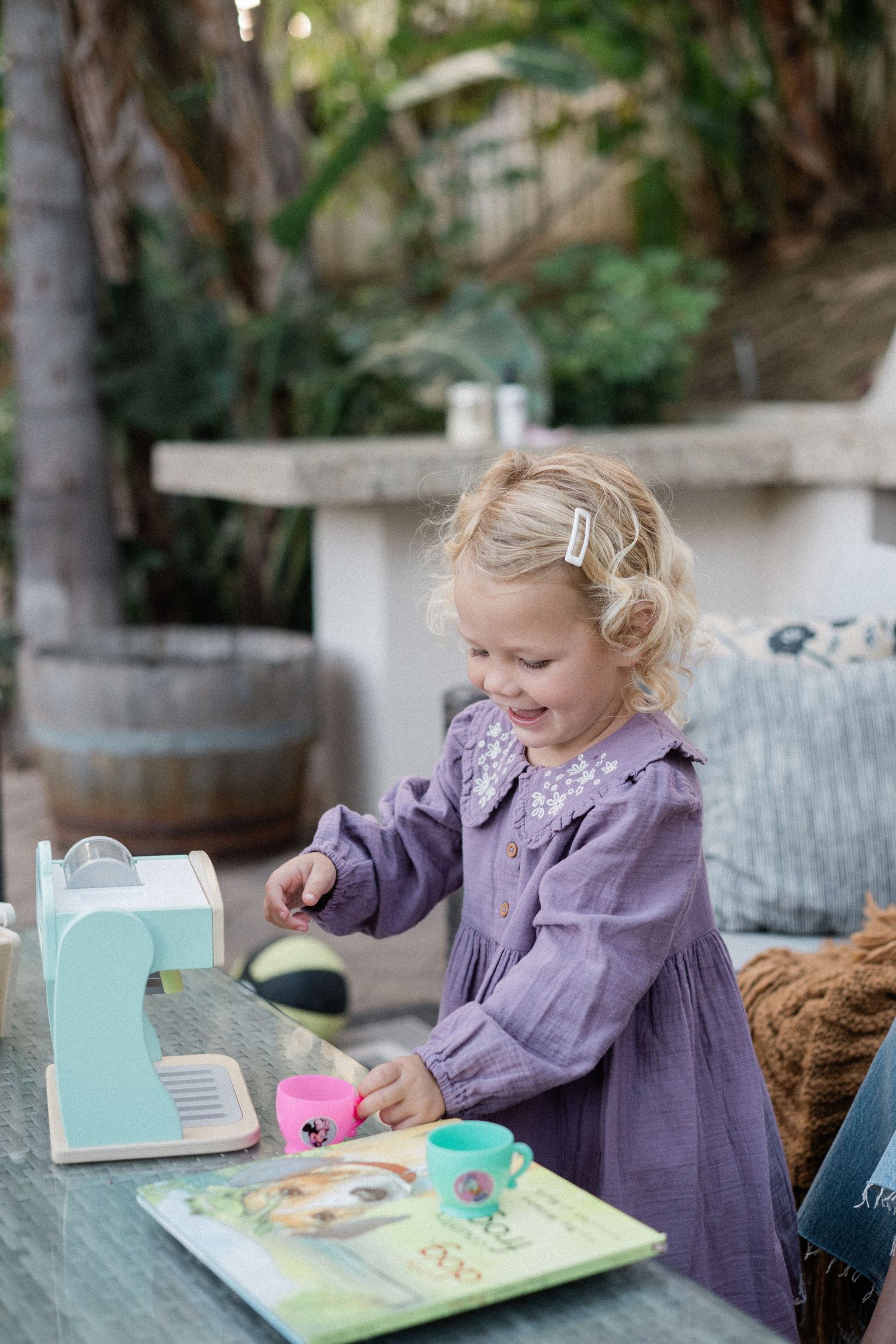 Blonde toddler girl in purple dress playing with a toy coffee maker and cups on an outdoor patio table.