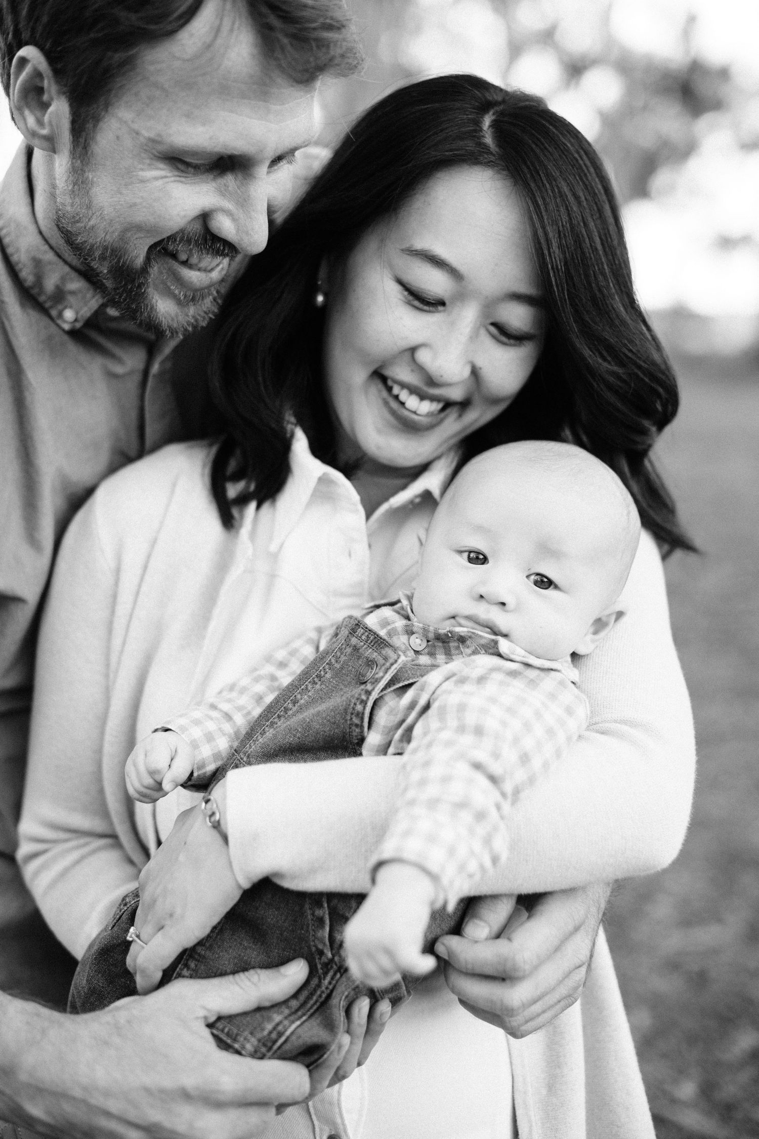 Smiling interracial couple holding their baby outdoors in a black and white family portrait photo.