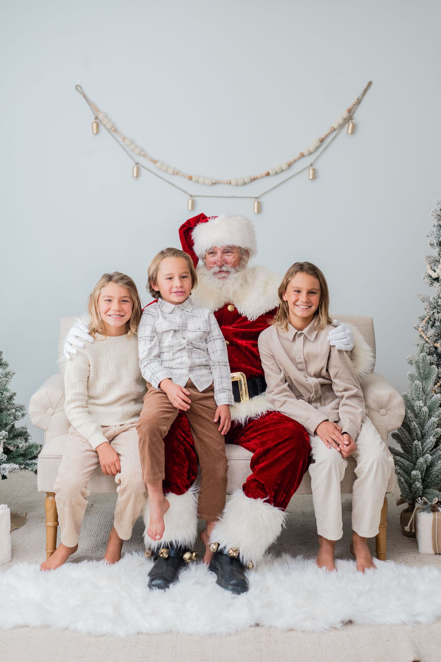 Three smiling children sitting with Santa Claus on a chair in a cozy Christmas-themed photo studio.
