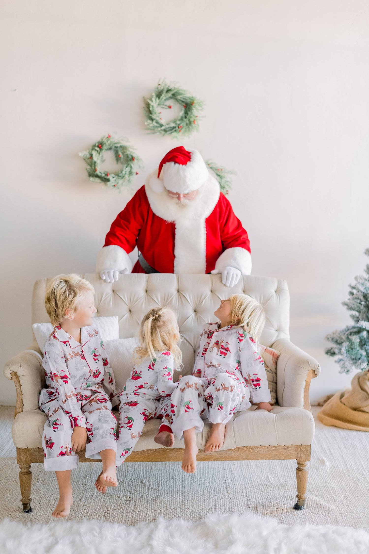 Three children in matching Christmas pajamas sit on a tufted sofa with Santa Claus in a festive holiday setting.