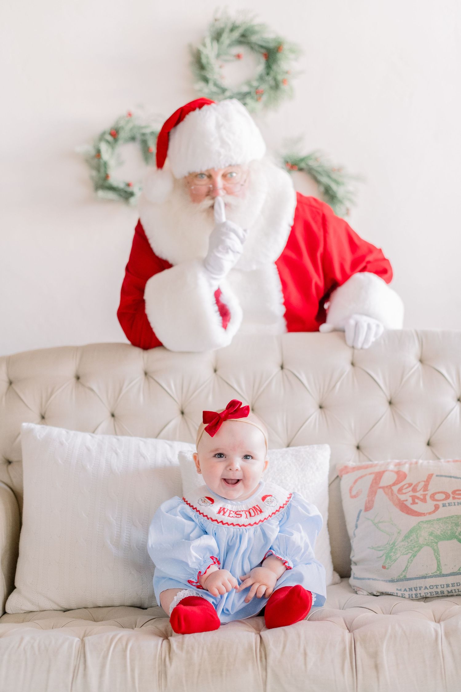 Baby girl in blue smocked outfit sits on tufted sofa while Santa Claus peeks over the back with a shush gesture.