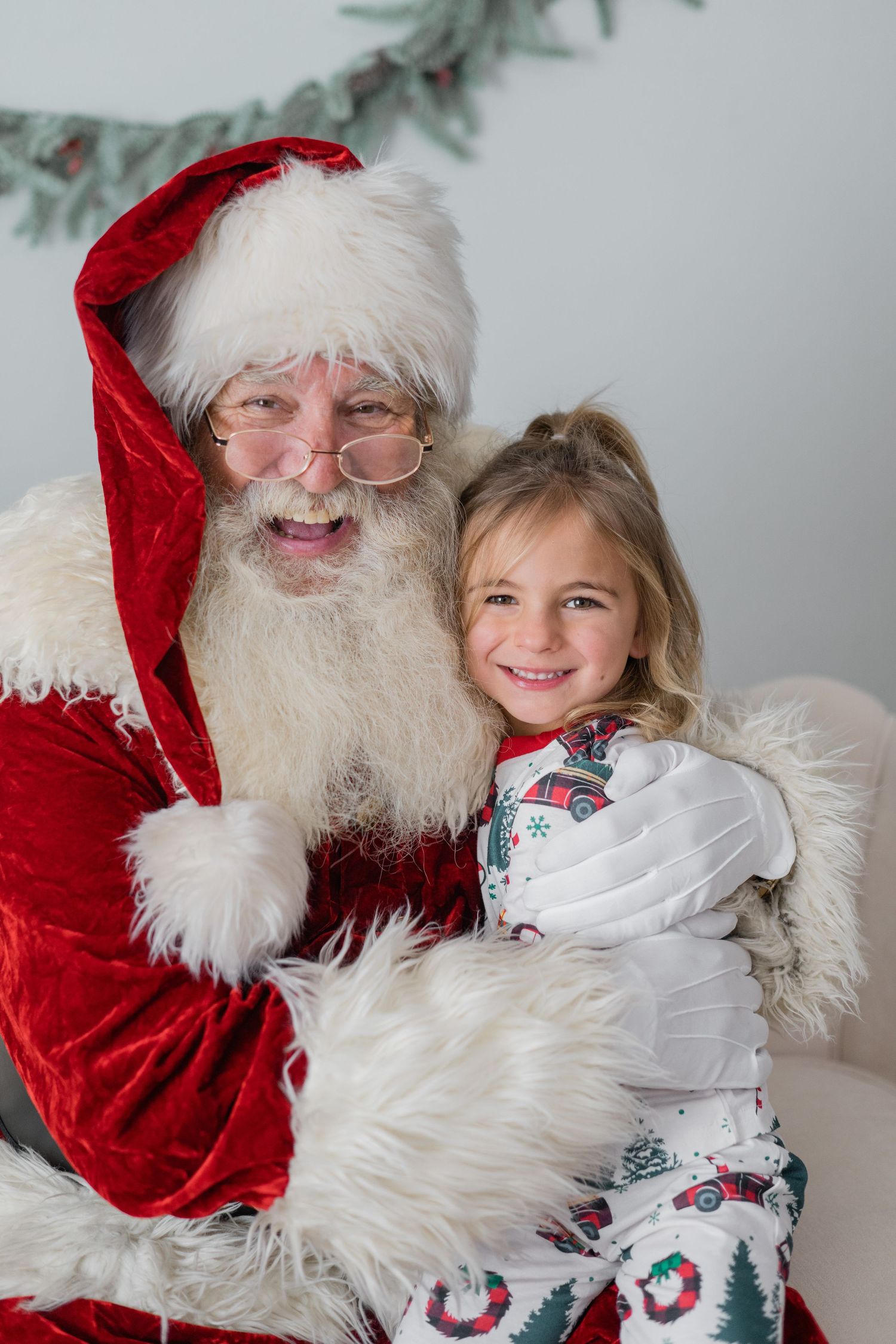 A smiling girl in Christmas pajamas sits with a laughing Santa Claus in a festive holiday setting.