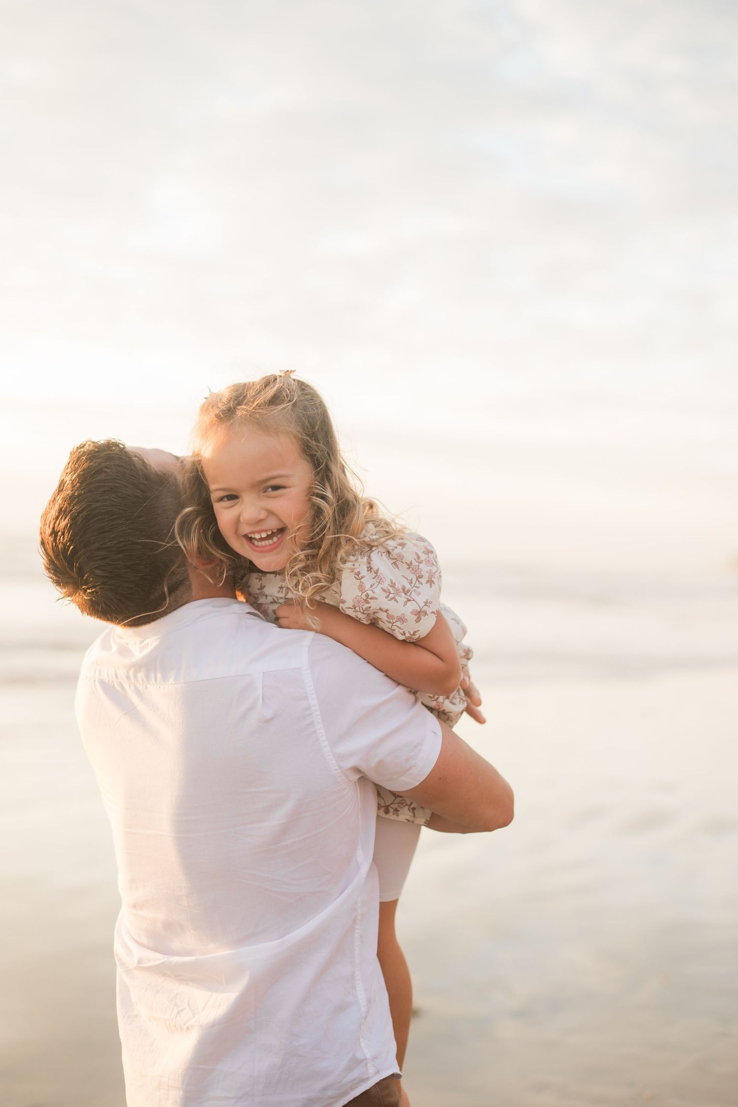 A joyful little girl laughs while being held by her father on a sunlit beach at sunset.