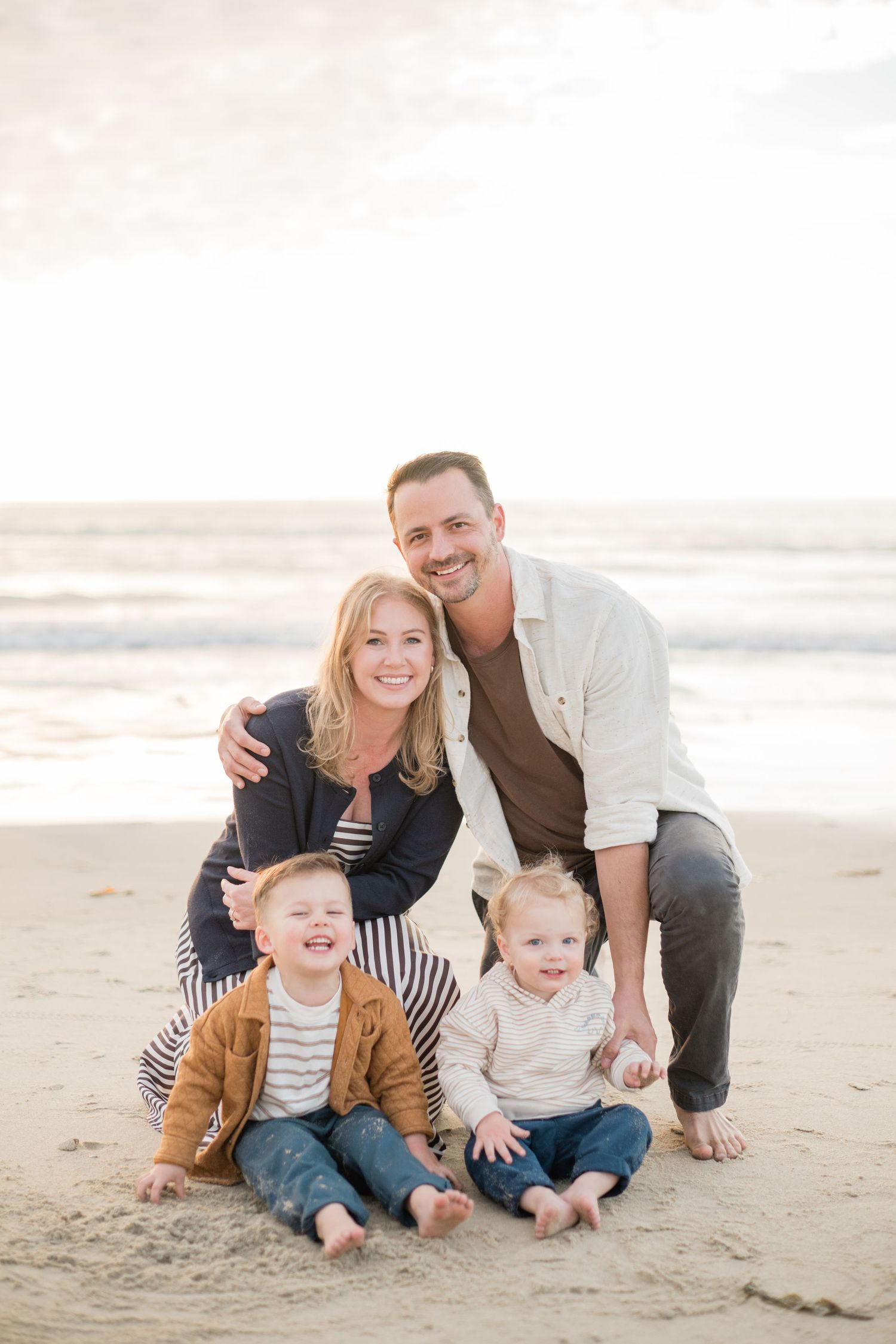 Happy family of four posing together on a sandy beach, with two young boys sitting in front of smiling parents.