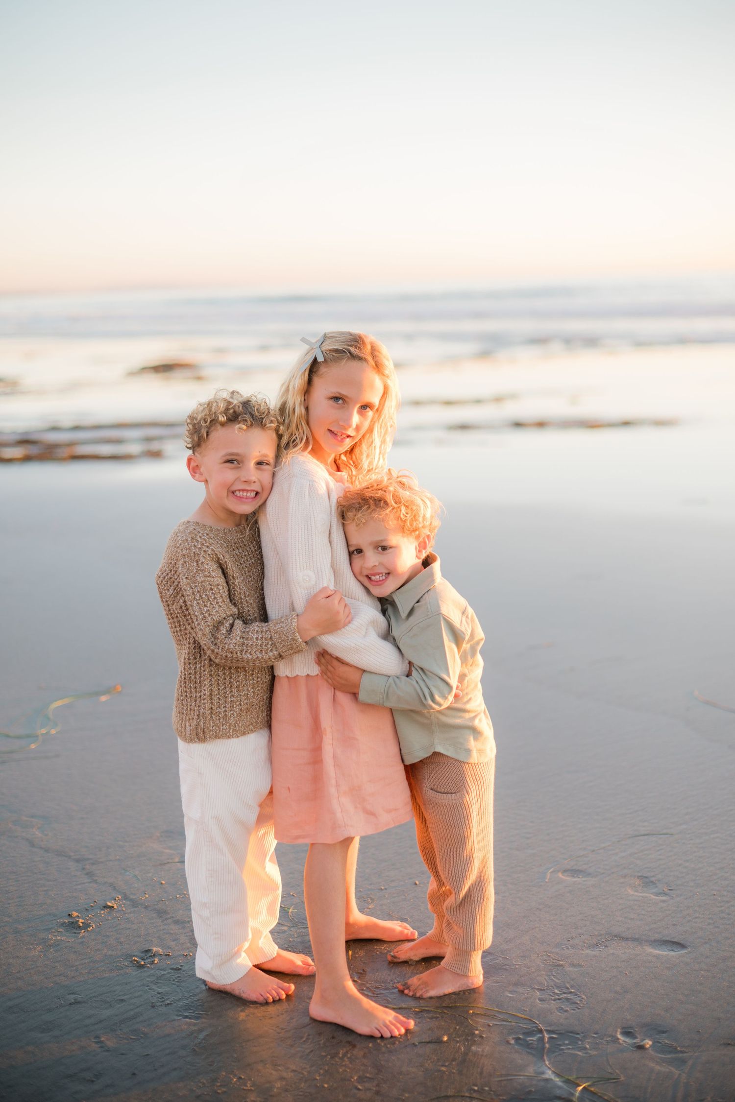 Three barefoot children hugging and smiling on a sandy beach at sunset with ocean waves in the background.