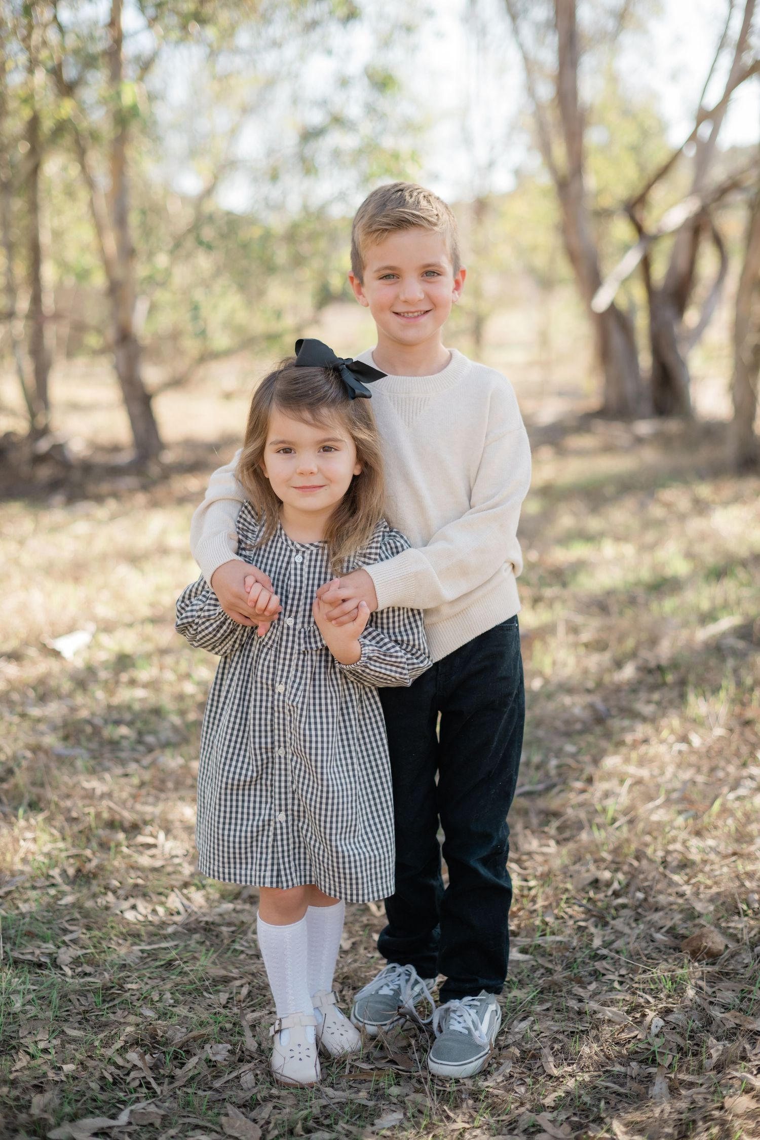 Two young siblings pose outdoors in a wooded area, girl in plaid dress with bow, boy in cream sweater and black pants.