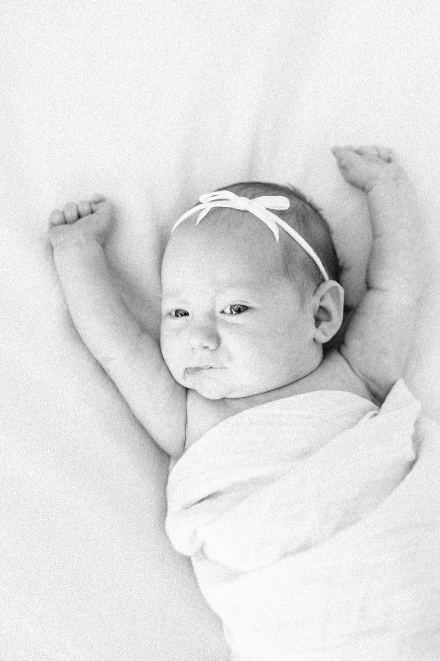 Black and white photo of a newborn baby girl with a bow headband, swaddled in white, arms raised.