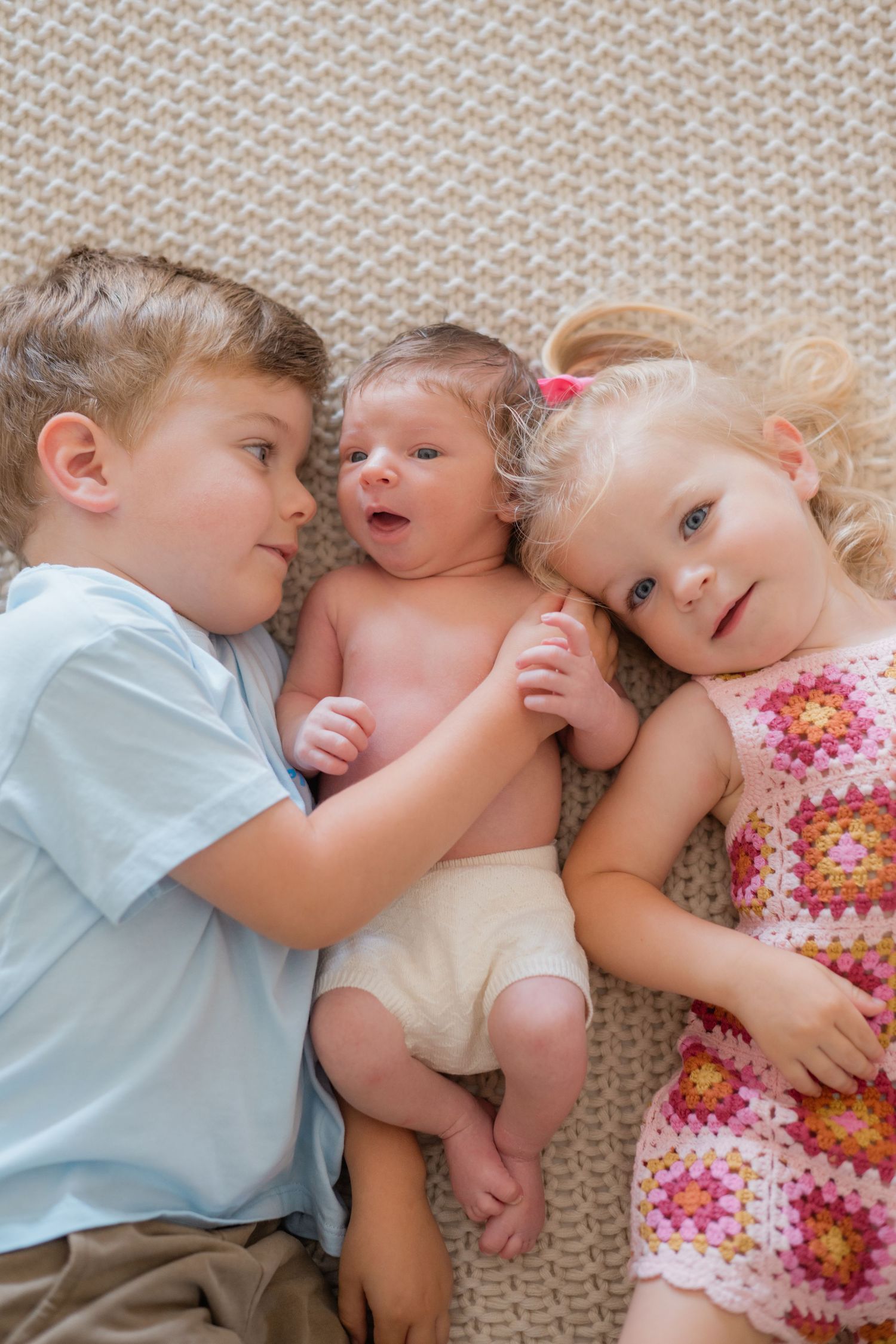 Two older siblings lying beside their newborn baby on a soft textured blanket.