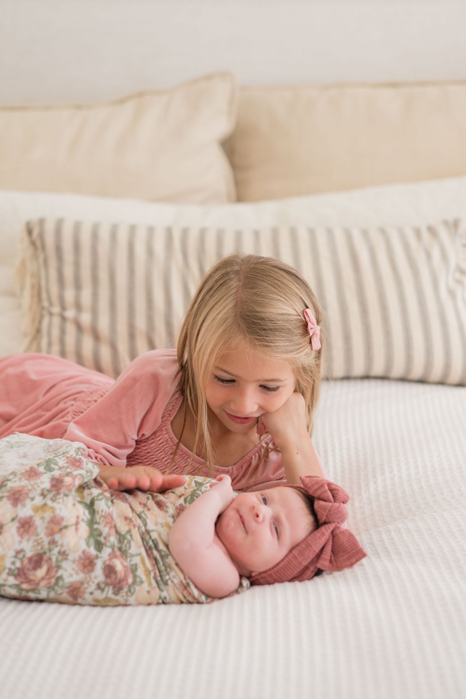 Young blonde girl smiling at swaddled newborn baby lying on white bed with striped pillows.
