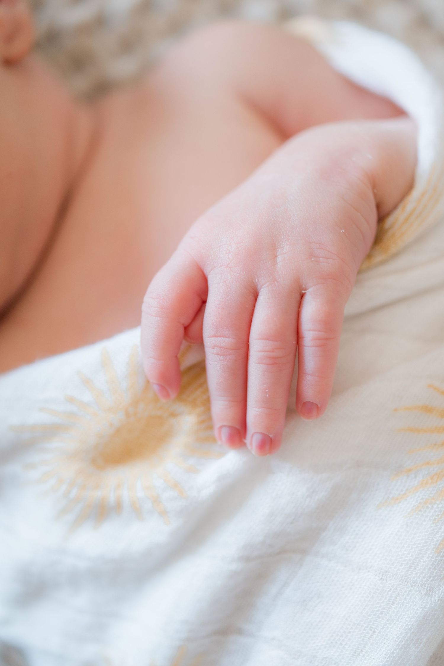 Delicate newborn baby hand with tiny fingers resting on a white blanket with golden sun print.