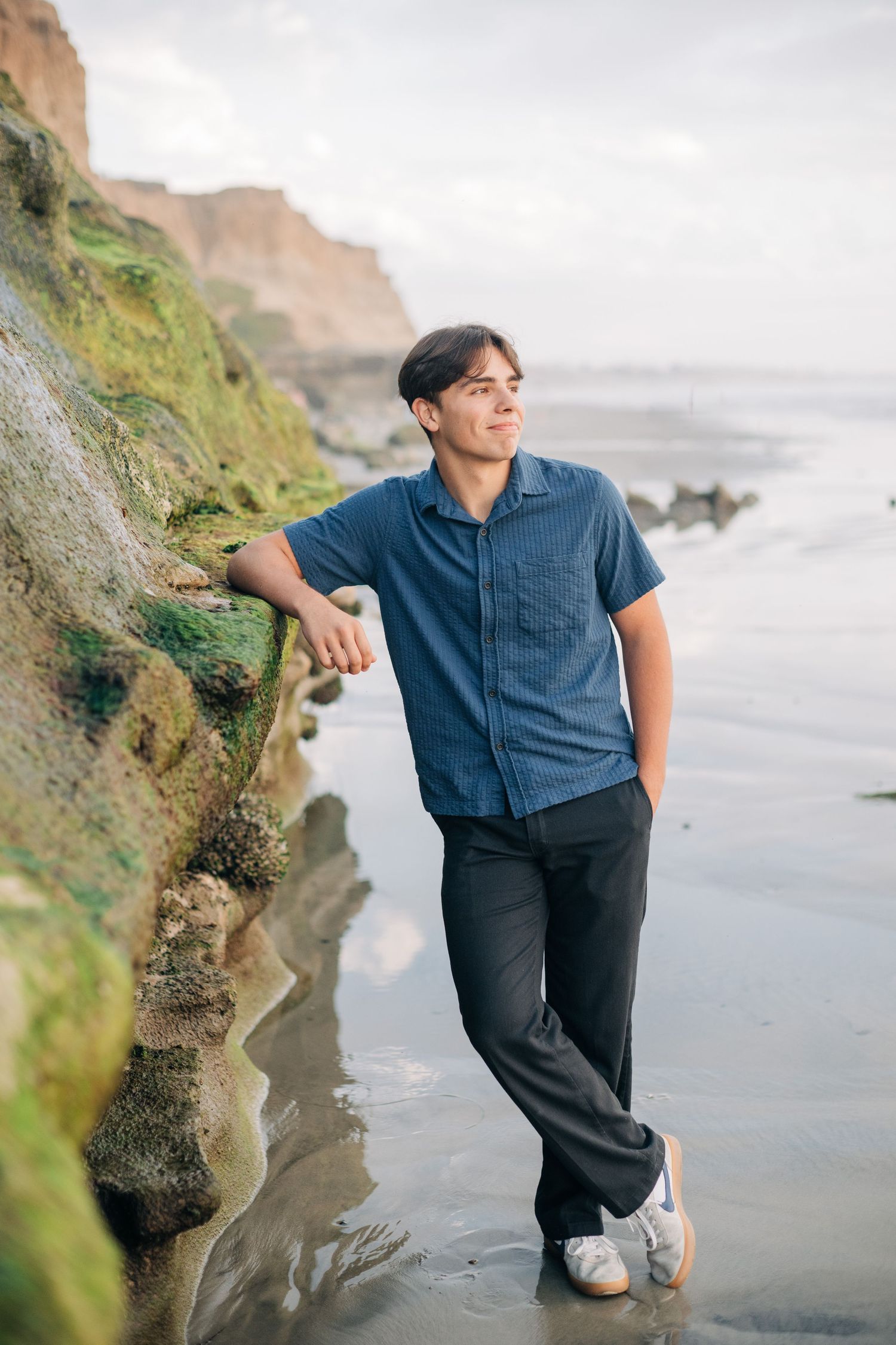 Young man in blue shirt leaning against mossy coastal rocks on a beach with cliffs in the background.