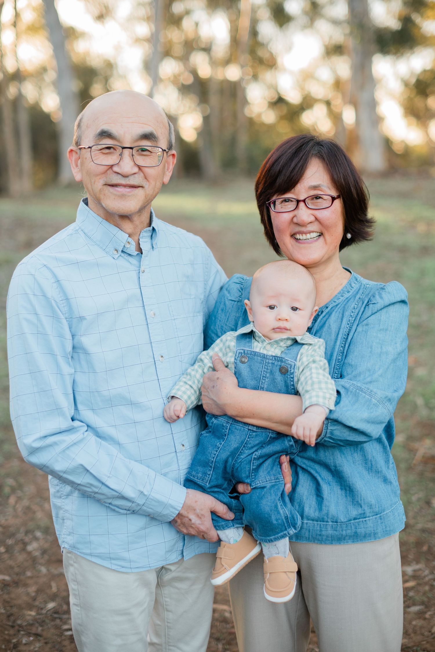 Grandparents with glasses smile proudly while holding an adorable baby in denim overalls outdoors.