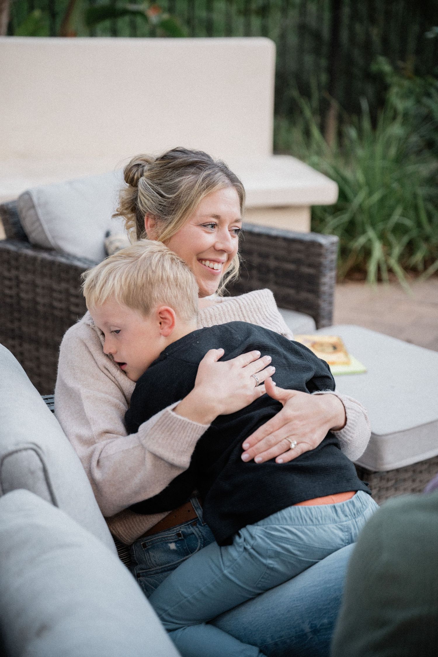 Smiling mother hugging her young blonde son on an outdoor patio sofa in a cozy backyard setting.