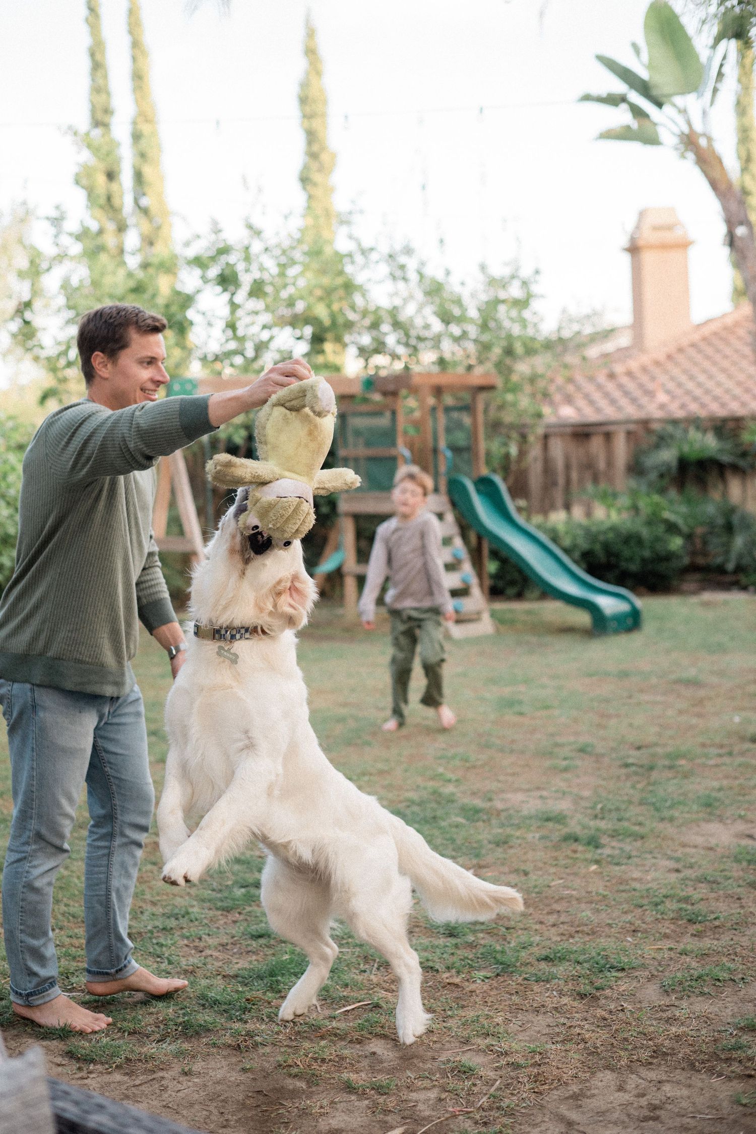 Man playing fetch with golden retriever in backyard while child plays near green slide.