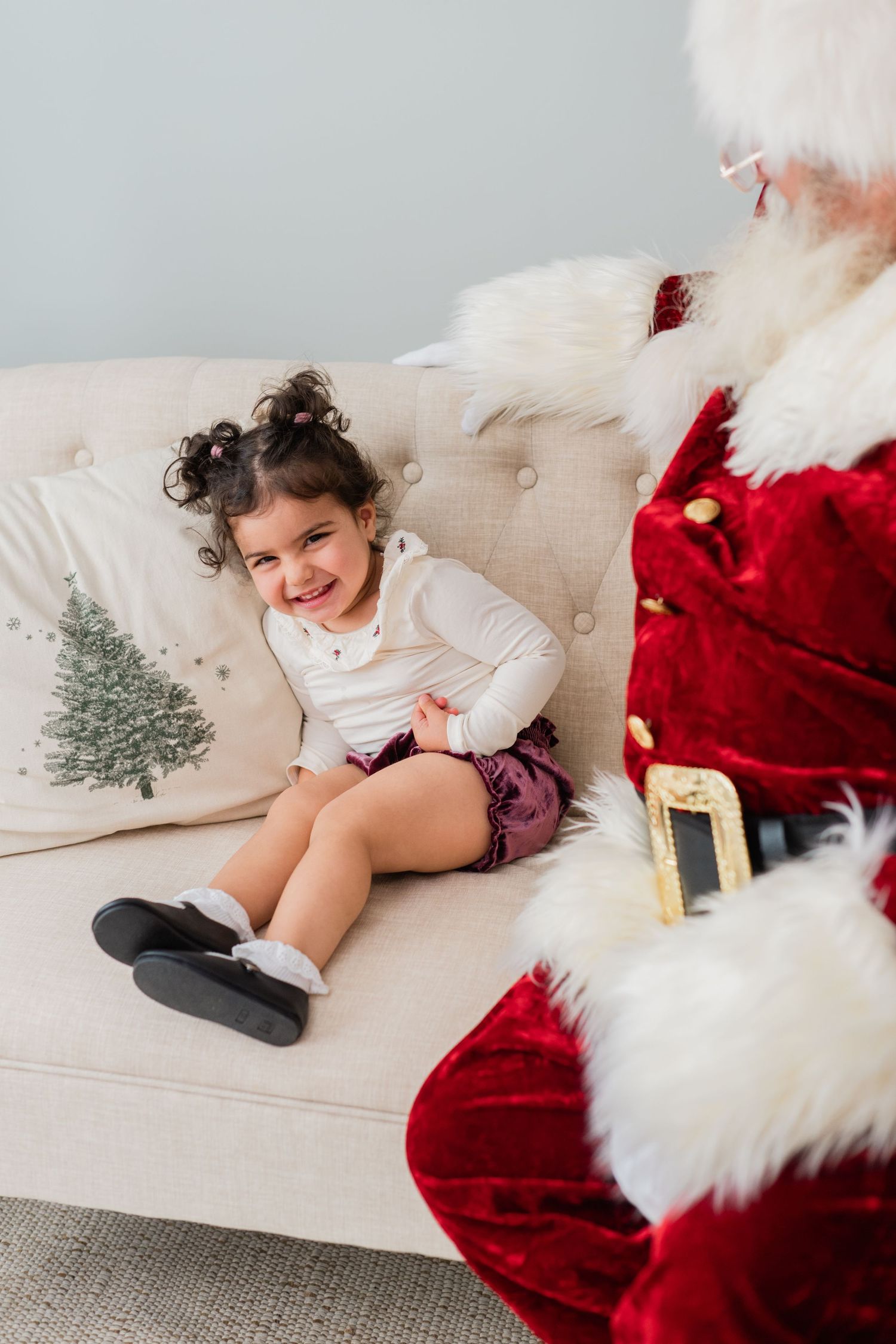 A laughing toddler girl in a plum skirt sits on a cream sofa next to Santa Claus during a Christmas photo session.