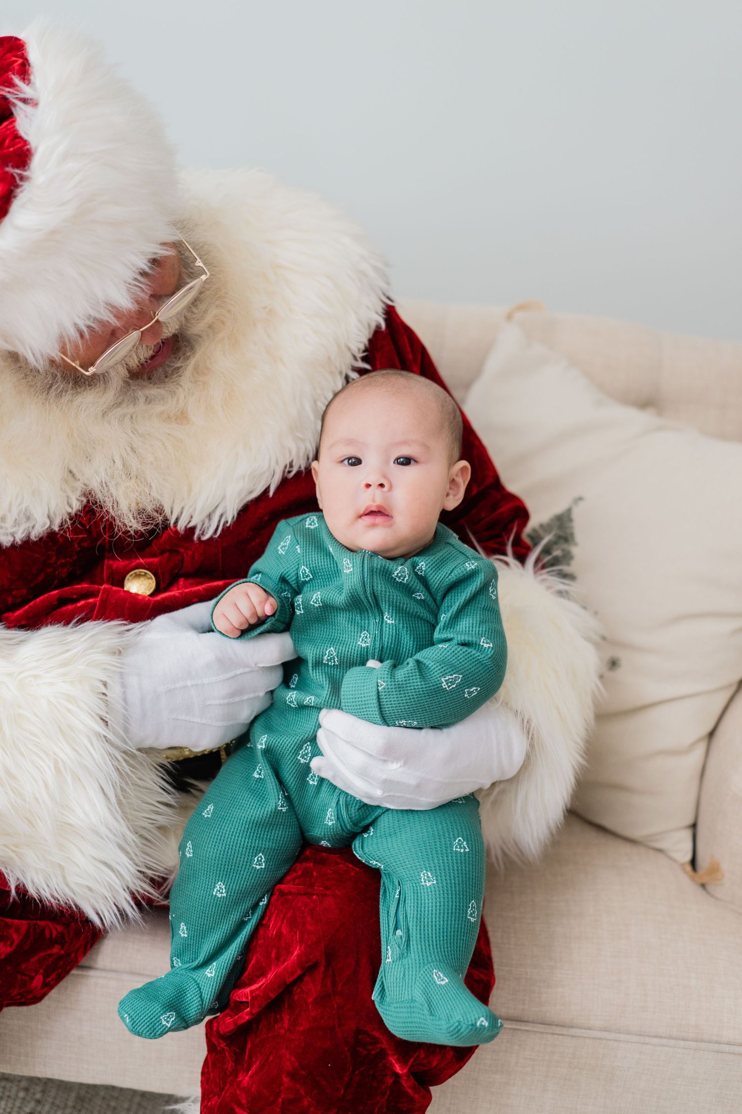 A baby in a green heart-print onesie rests in Santa Claus's white-gloved hands on a cream couch.