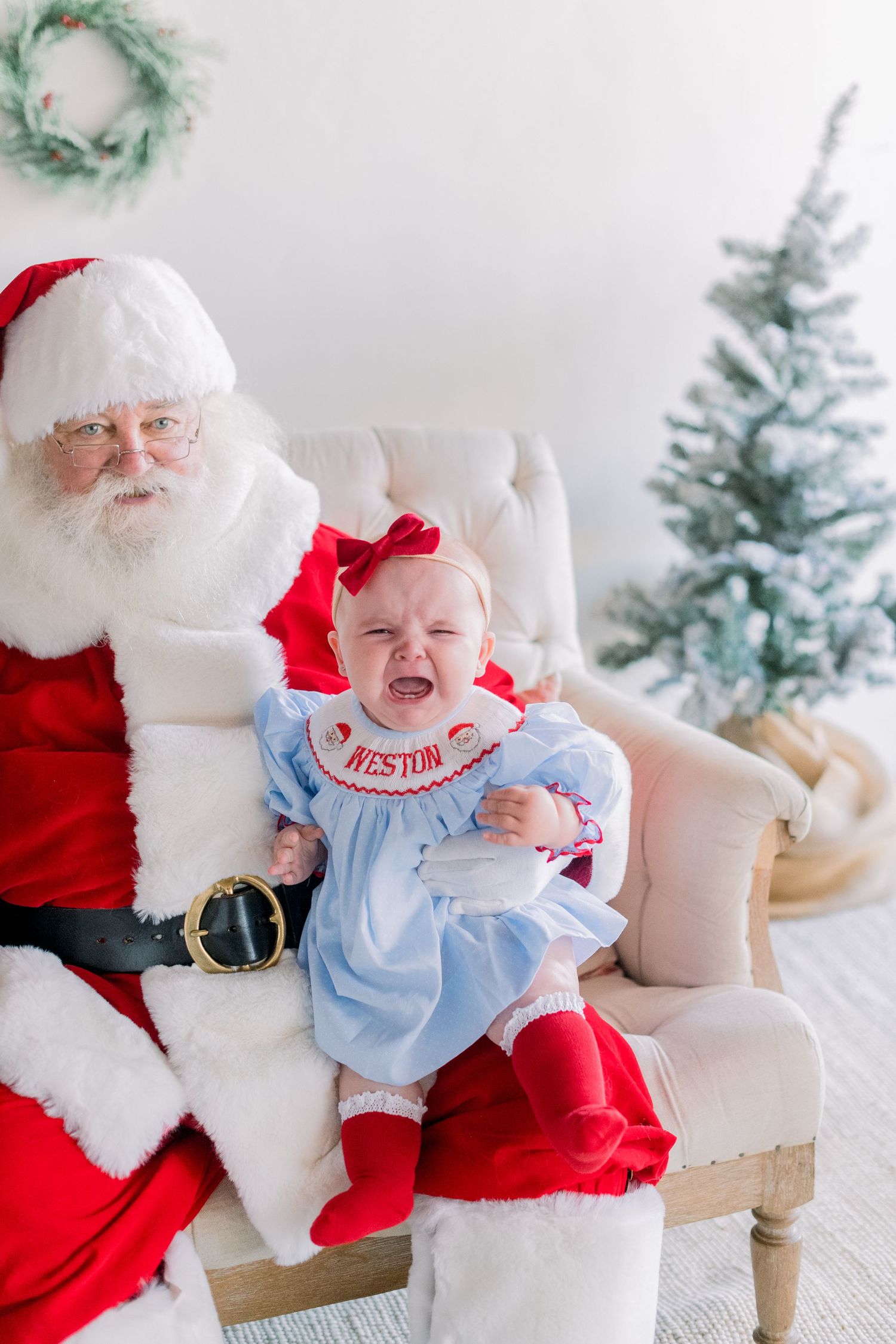 Crying baby named Weston in blue dress sitting on Santa Claus lap during Christmas photo session near decorated tree.
