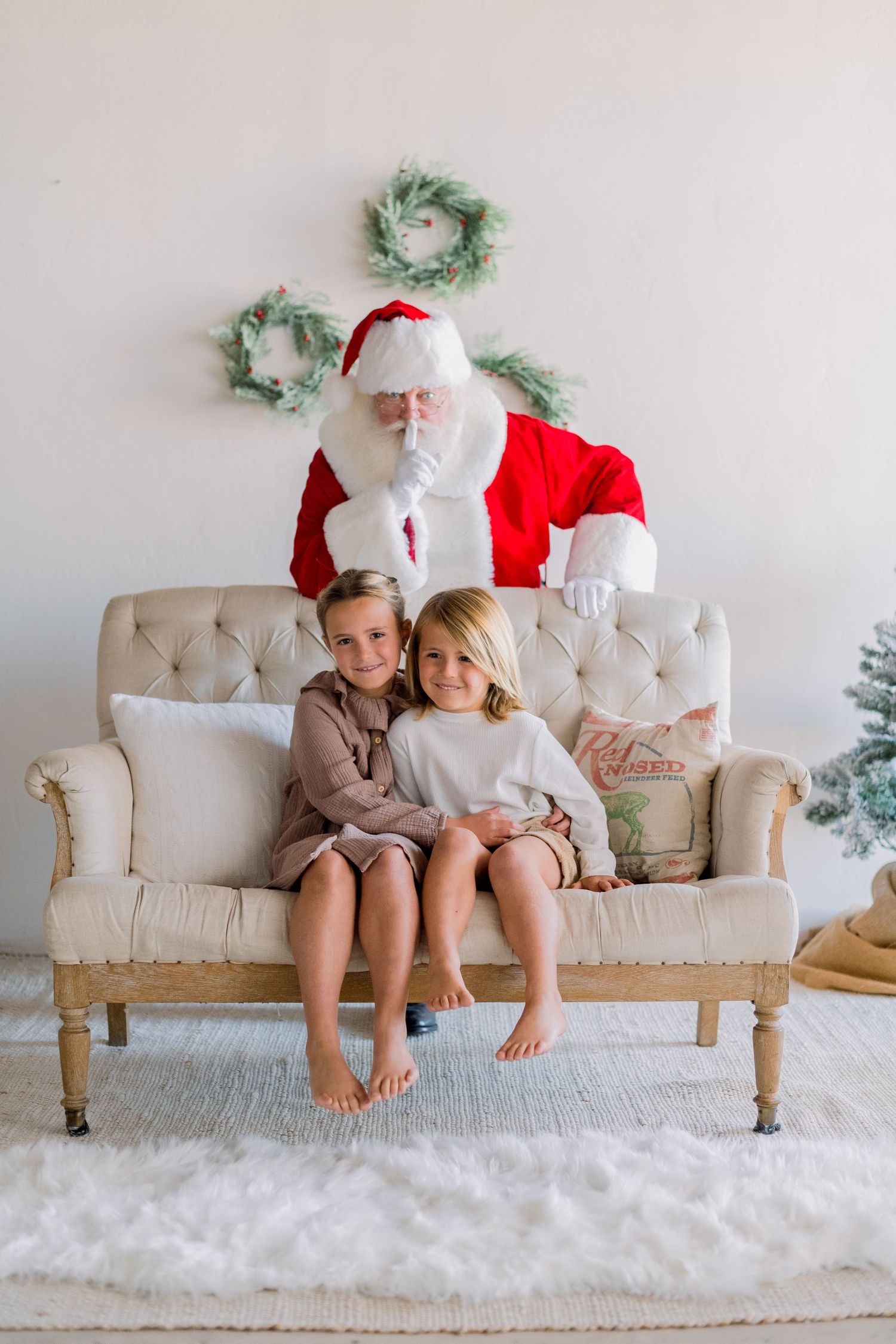 Two young girls sit together on a cream tufted sofa with Santa Claus behind them in a Christmas setting.