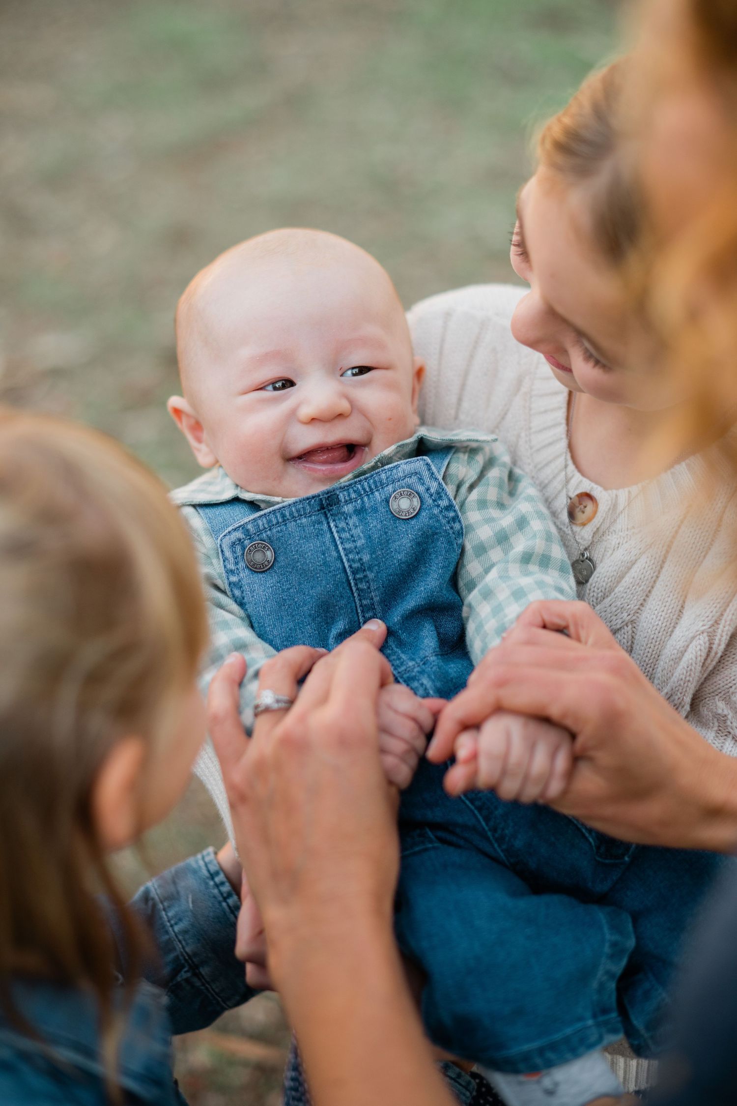 A smiling baby in denim overalls is held by a woman while a child reaches out to play.
