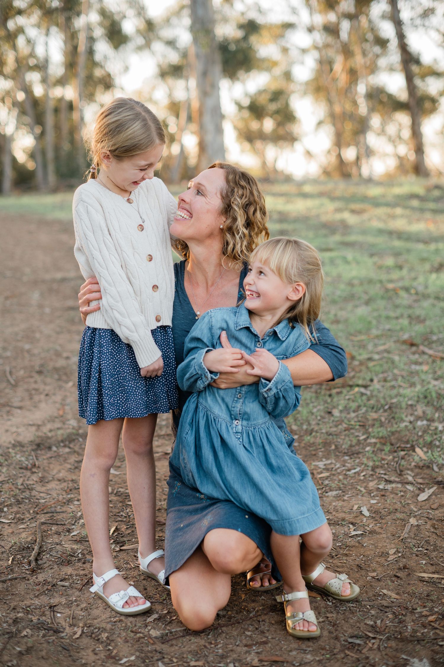 A laughing mother kneels in a park hugging her two young blonde daughters wearing blue skirts and white tops.