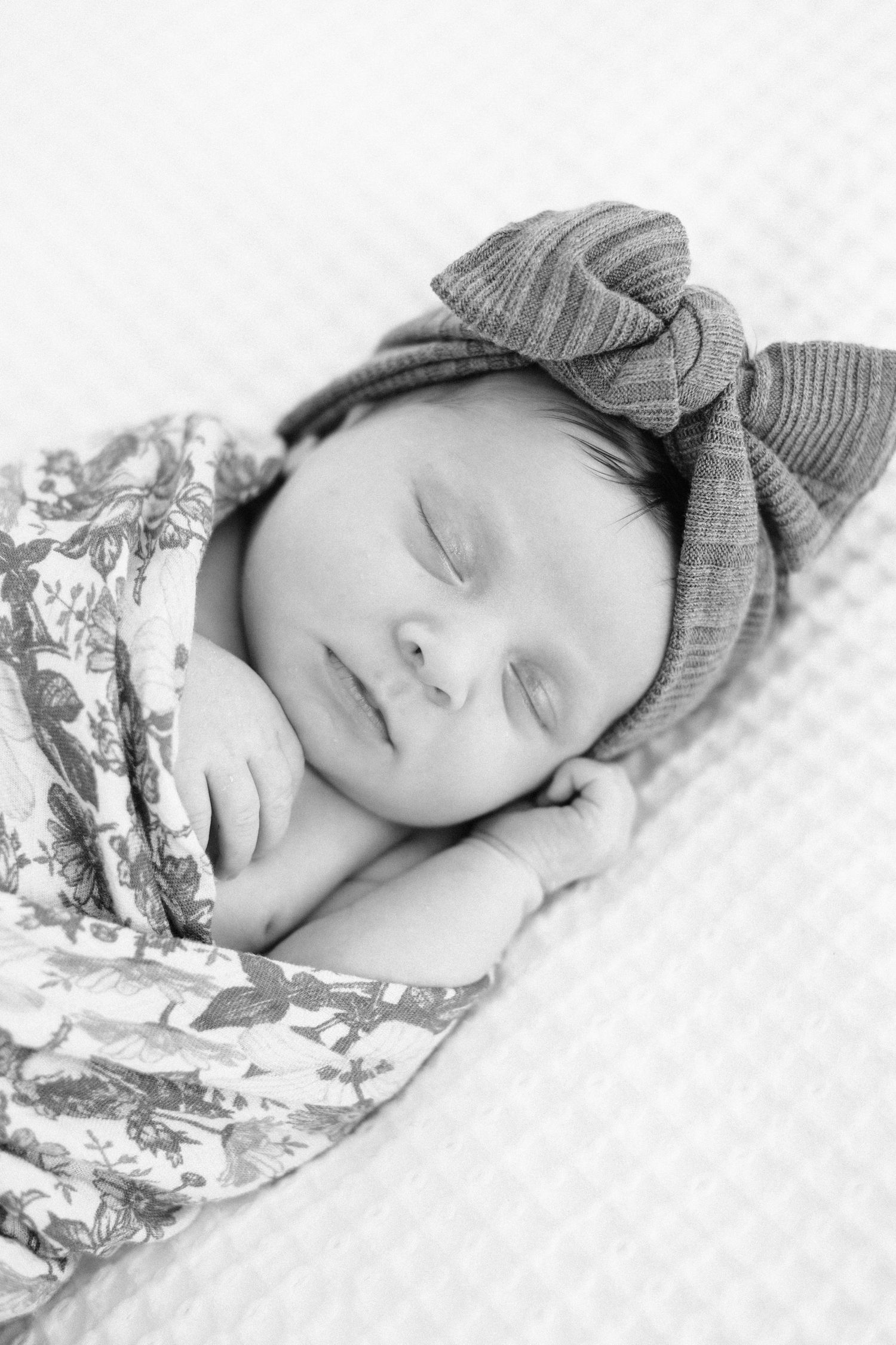 Black and white photo of a sleeping newborn baby wrapped in a floral swaddle wearing a knit bow headband.