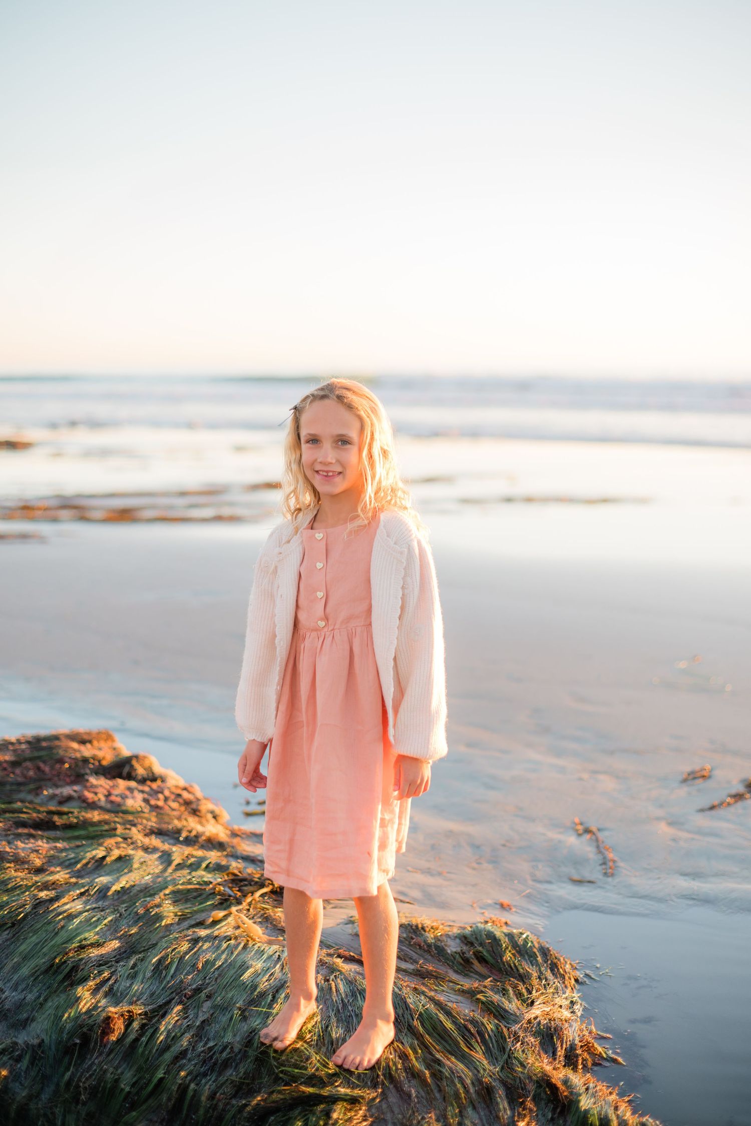 Young blonde girl in pink dress and cardigan standing barefoot on mossy rocks at a serene beach during golden hour.