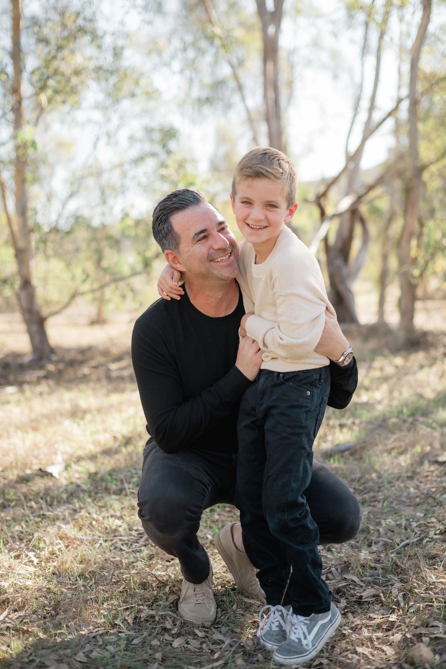 A man crouching beside a smiling young boy outdoors among trees, sharing a warm embrace in natural light.