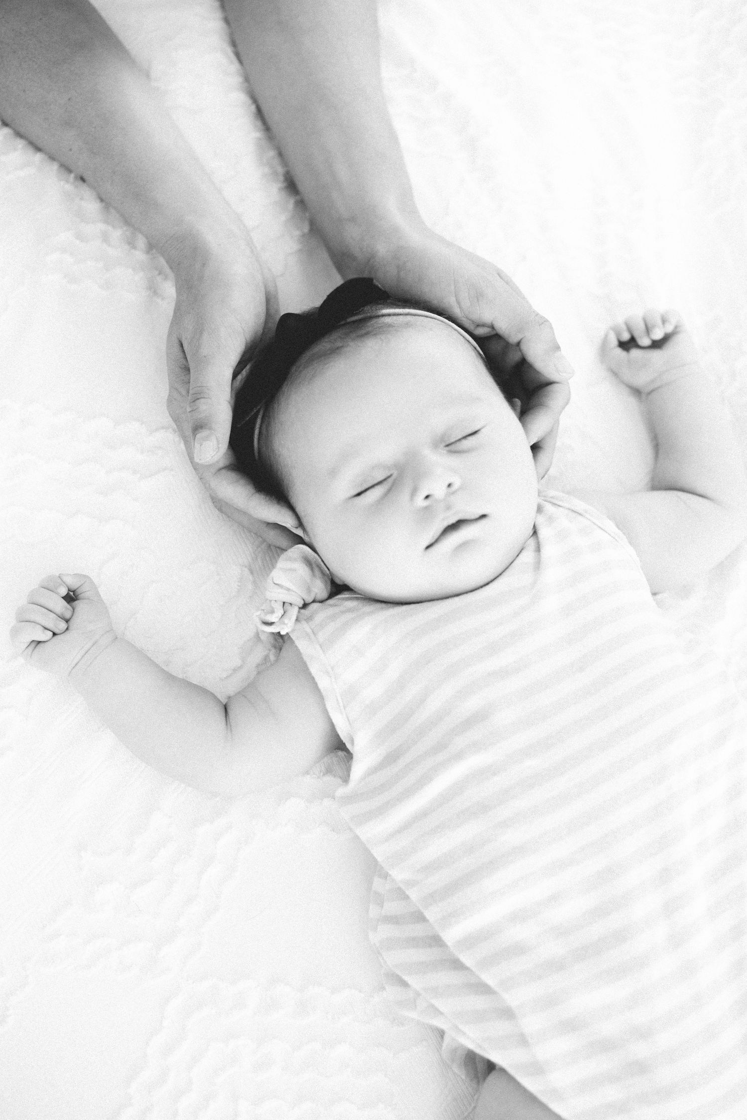 Black and white photo of a sleeping newborn baby in striped onesie with adult hands cradling its head.