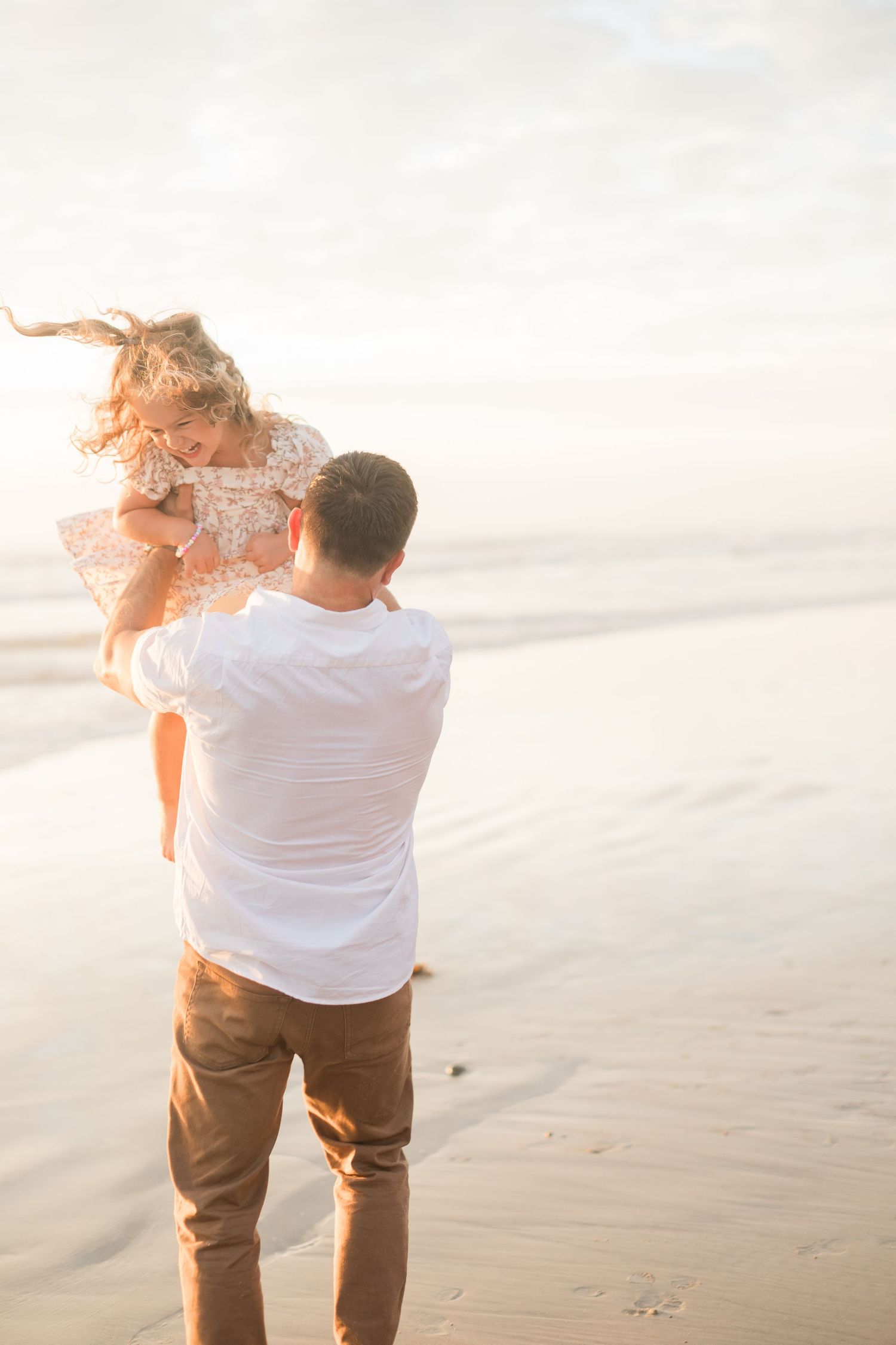 Father joyfully lifts his laughing daughter into the air on a golden sunlit beach during a family photoshoot.