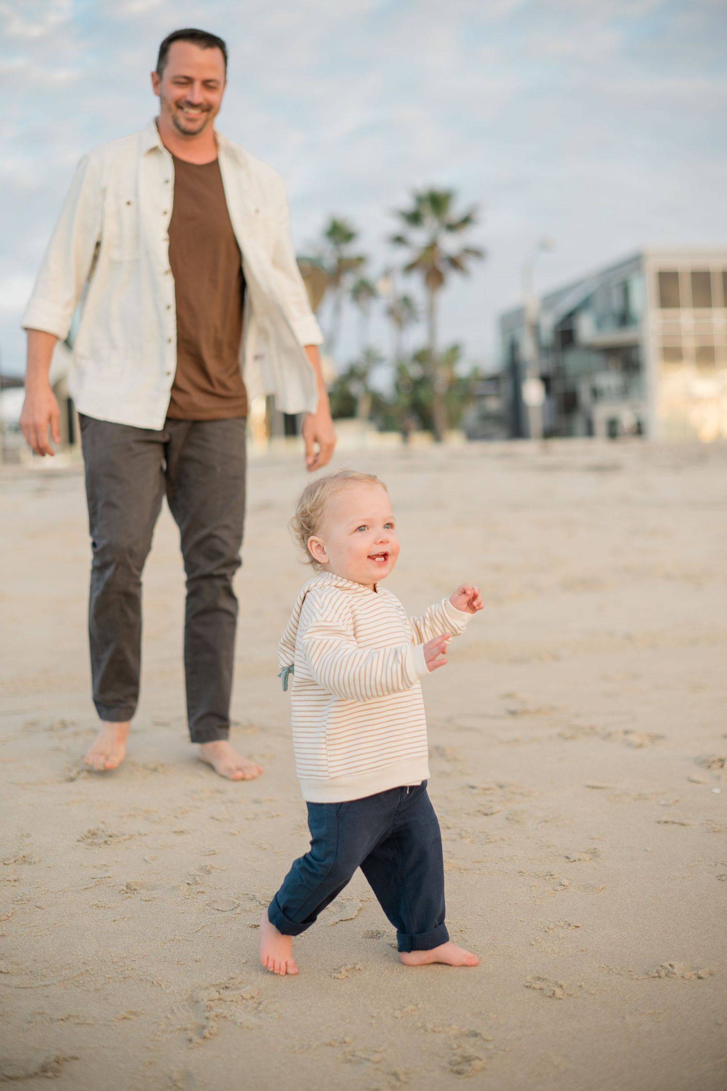 A happy toddler takes first steps on a sandy beach while a smiling father walks closely behind near palm trees.