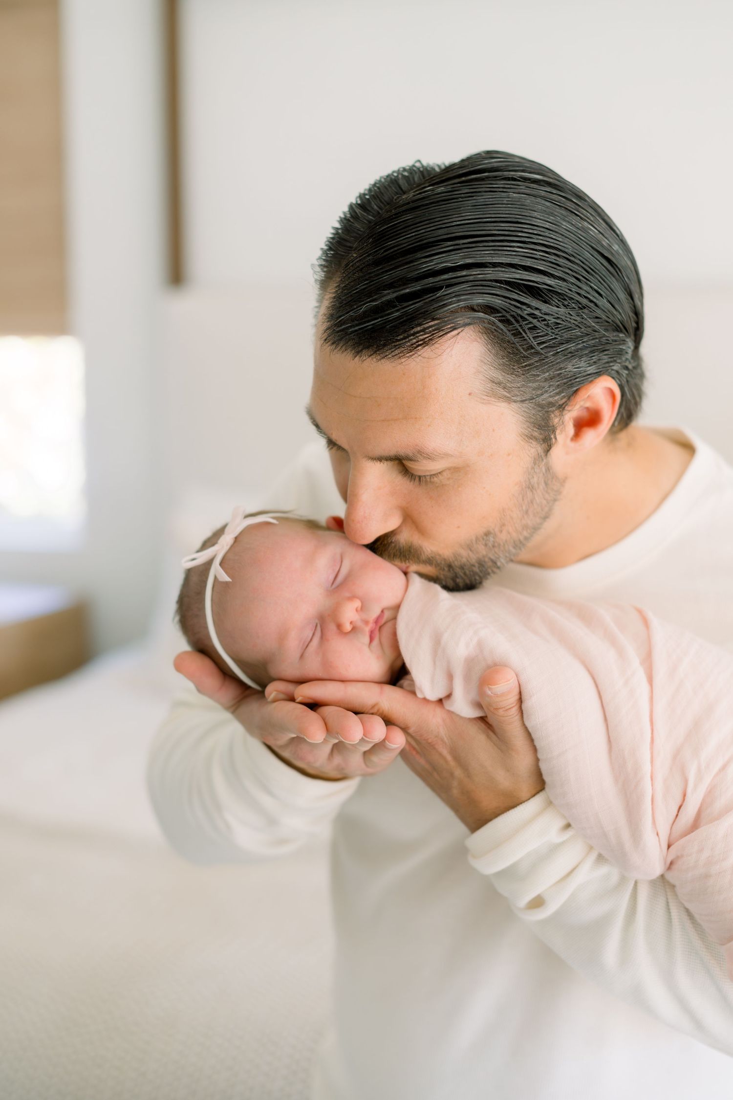 A father tenderly kisses his sleeping newborn baby, cradling the infant gently in his hands against a soft white background.