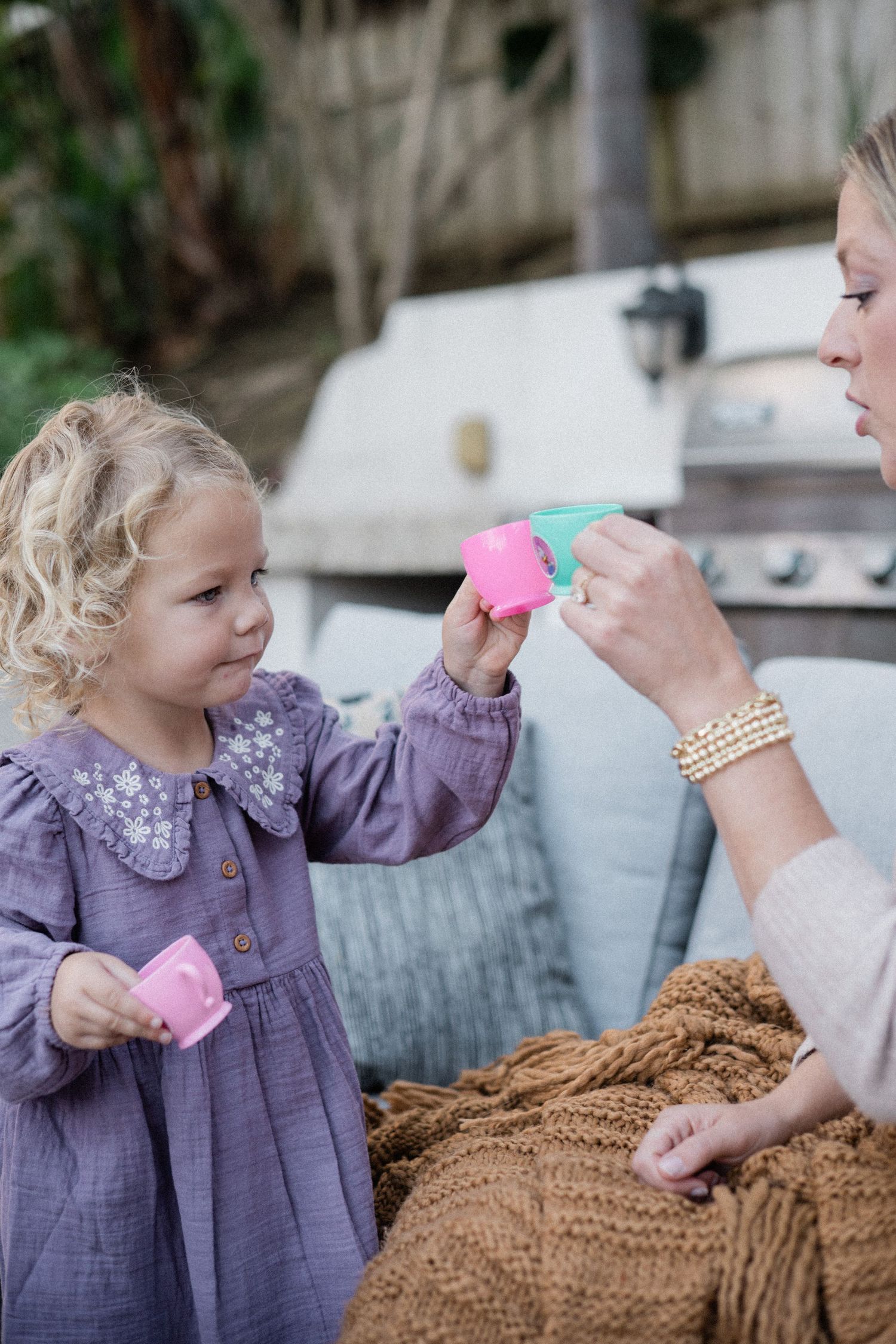 A young girl in a purple dress plays tea party with her mother outdoors, sharing colorful toy cups.