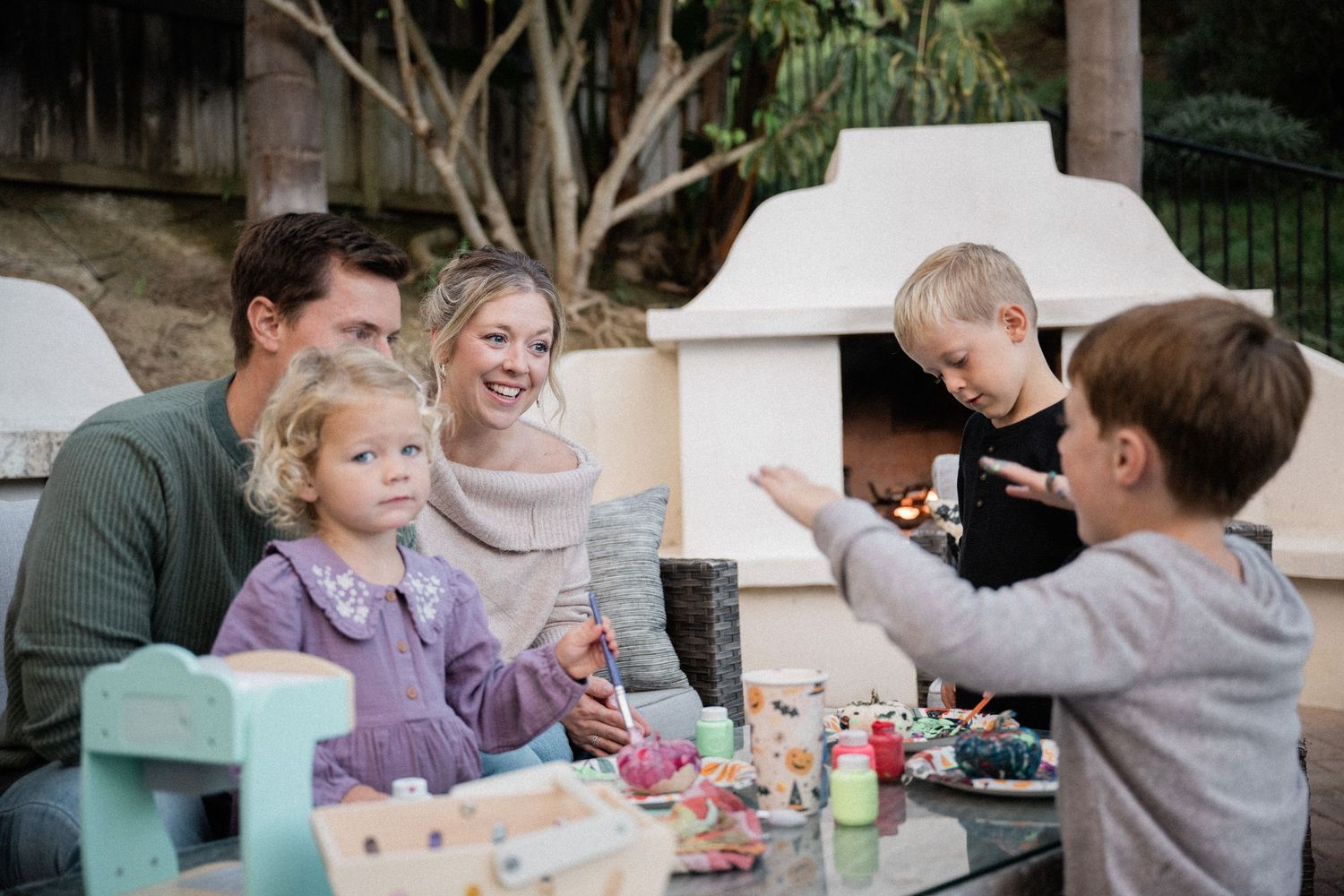 Family gathered outdoors around a table, enjoying playtime near a stone fireplace in a cozy backyard setting.