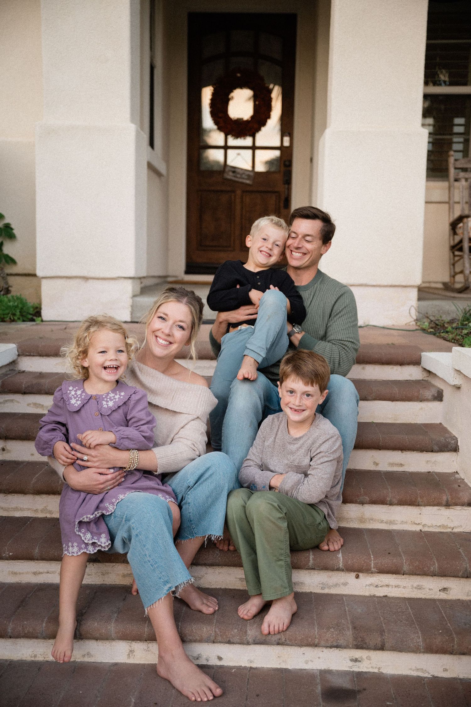 Happy family of five sitting barefoot on front porch steps, smiling and laughing together outside their home.