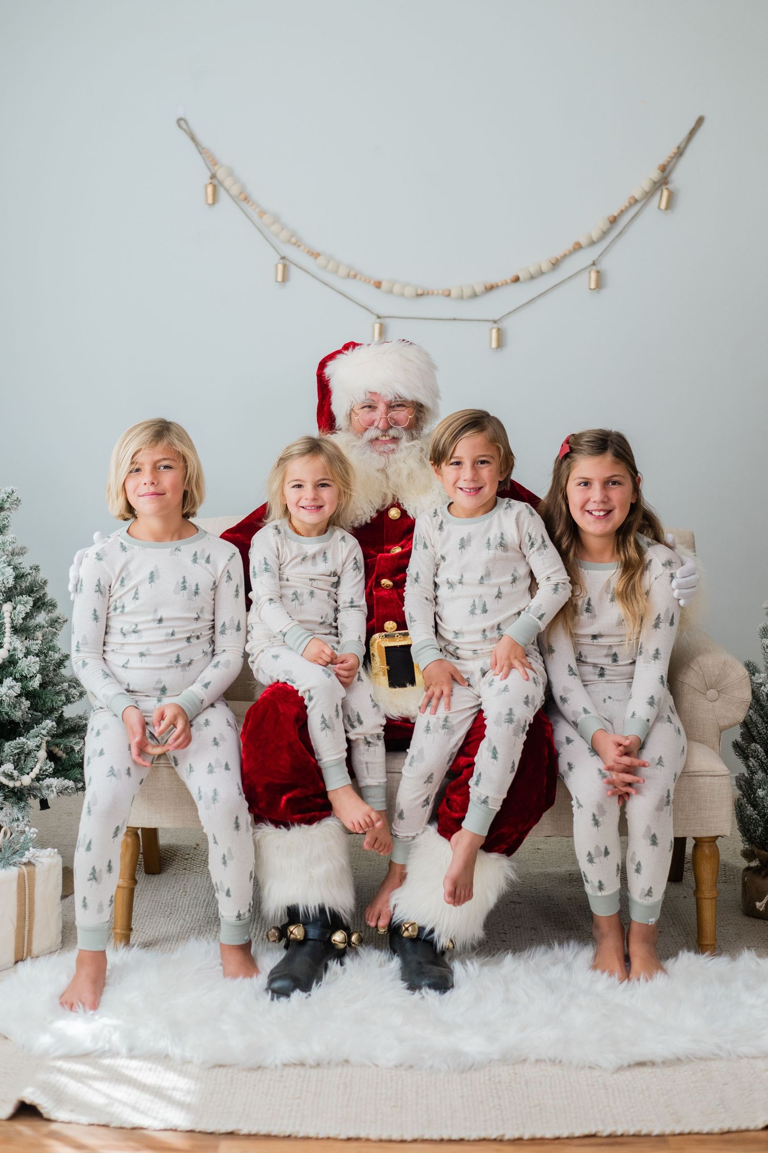 Four children in matching Christmas tree pajamas smile with Santa Claus on a cream couch with holiday decorations.
