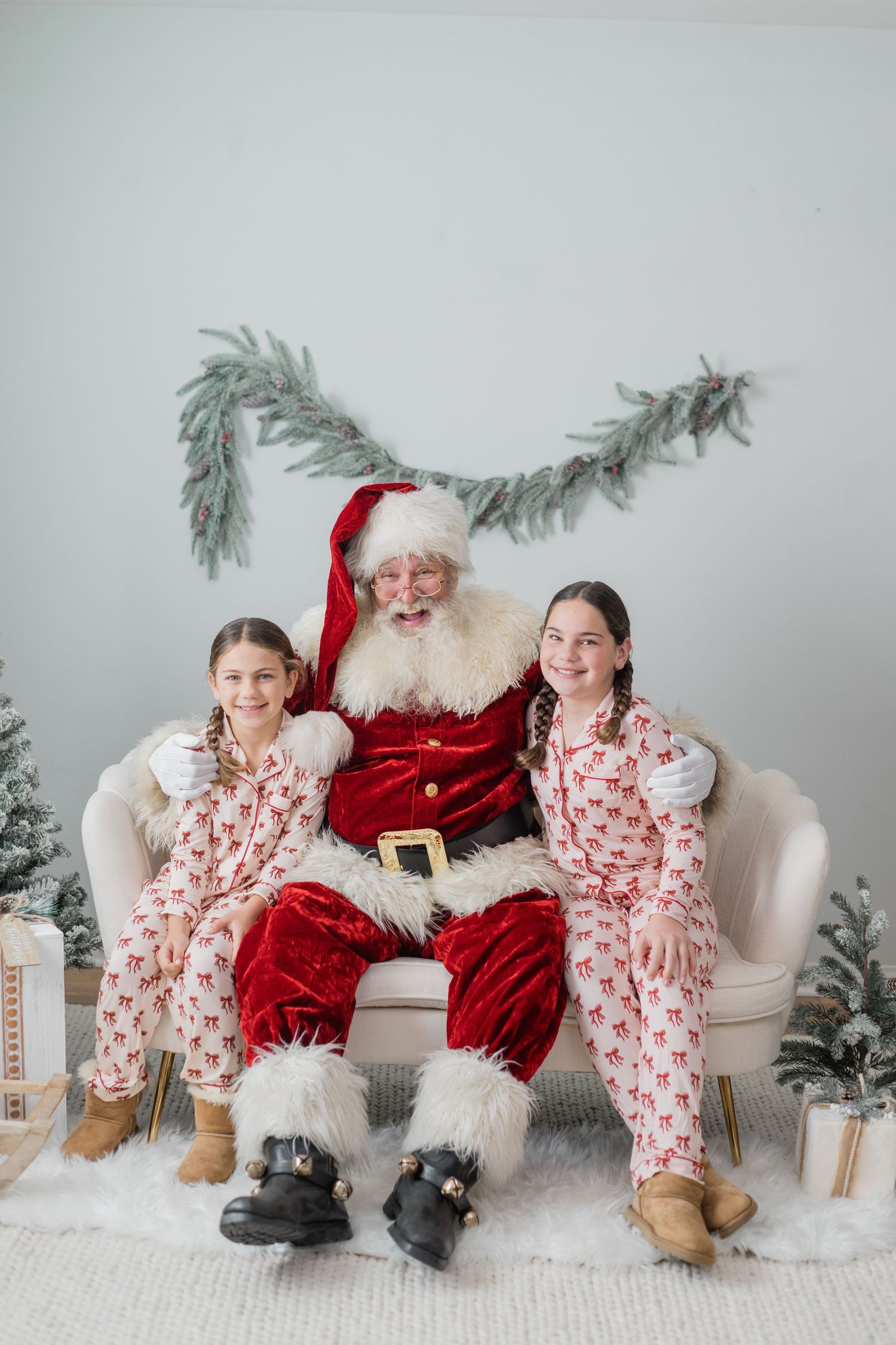 Two girls in matching reindeer pajamas sit with Santa Claus on a couch in a Christmas decorated room.