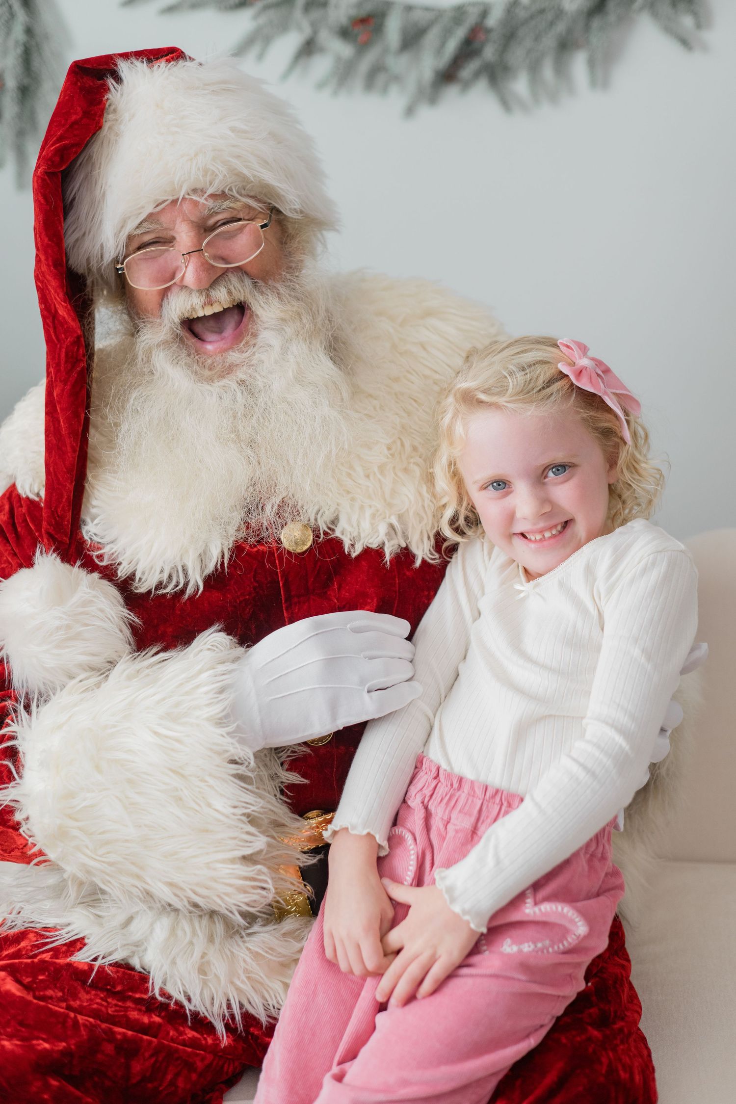 A laughing Santa Claus poses with a smiling young girl in pink on a white couch with Christmas decor.