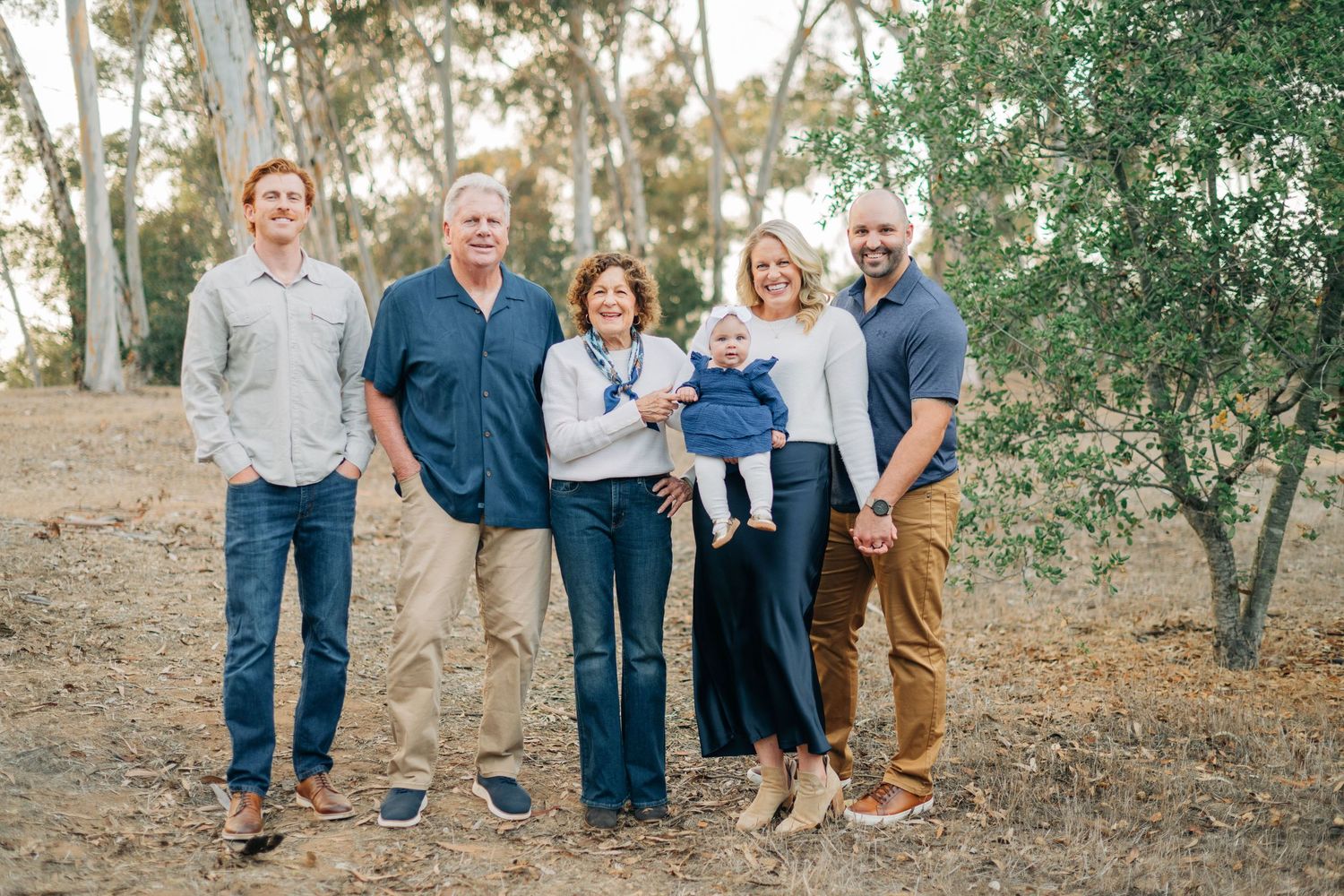 Family of six poses outdoors among eucalyptus trees, including grandparents, adult children, and a baby.
