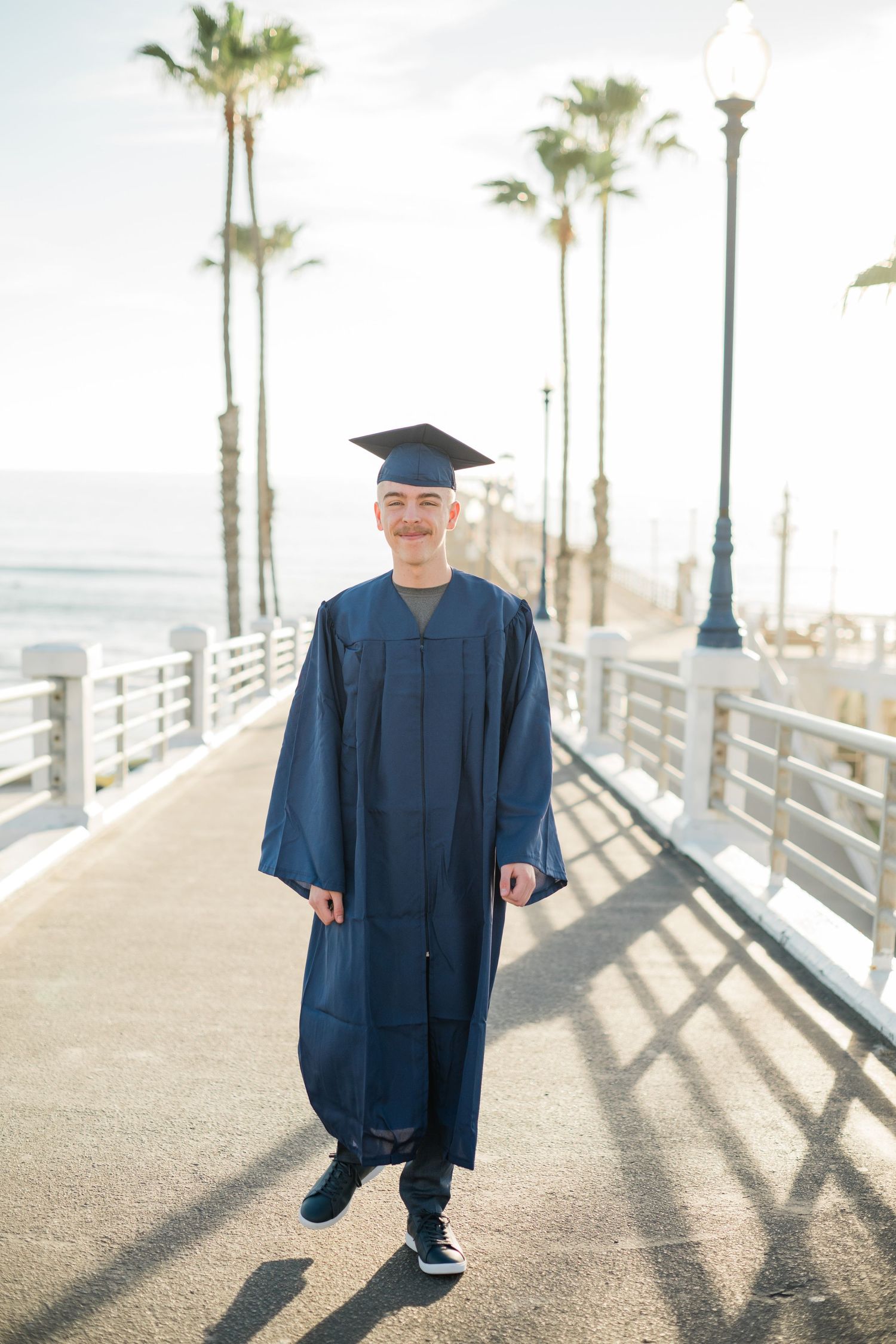 Young man in blue graduation cap and gown smiling on a sunny boardwalk with palm trees and ocean in background.