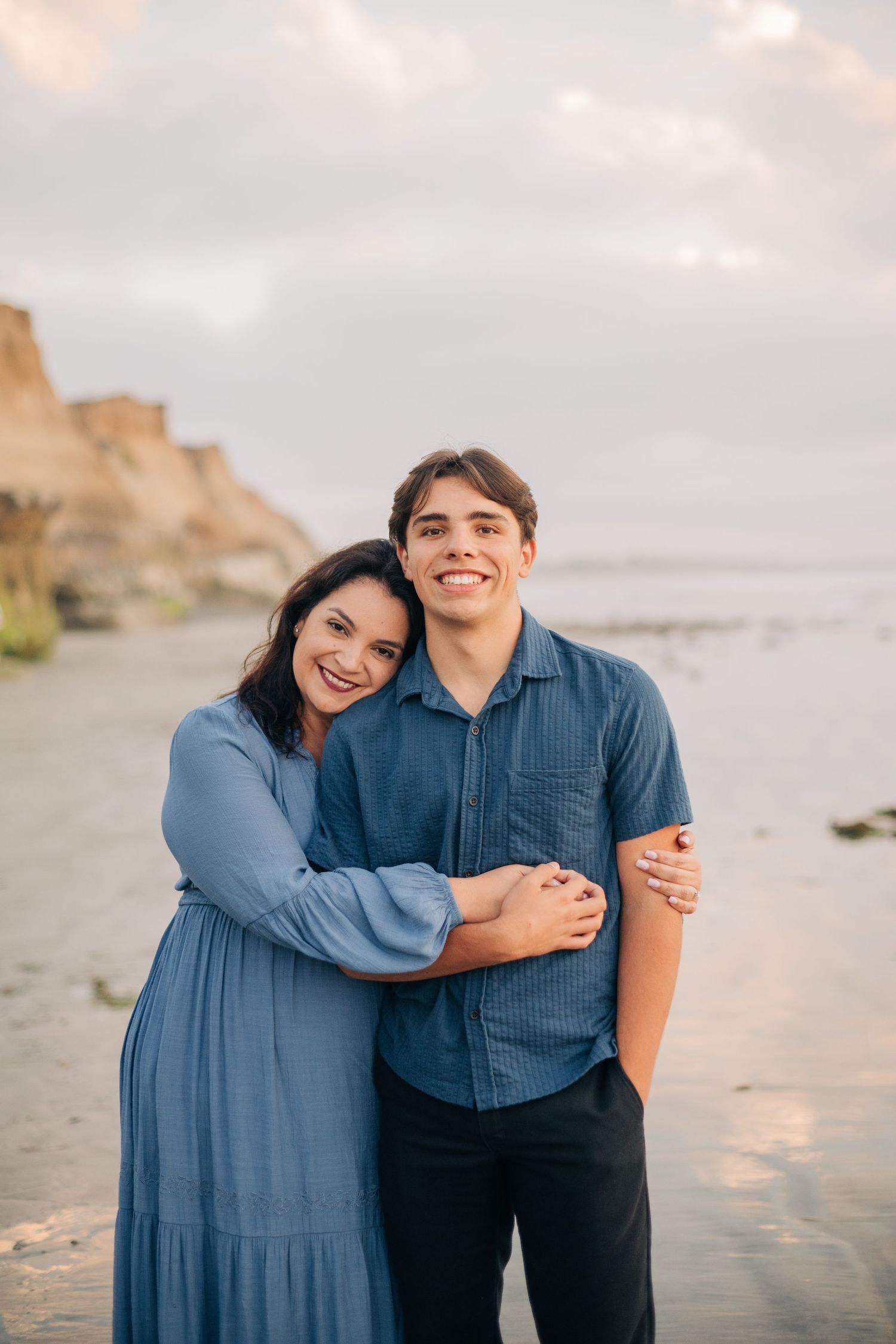 A smiling couple in matching blue outfits embracing on a sandy beach with cliffs and ocean in the background.