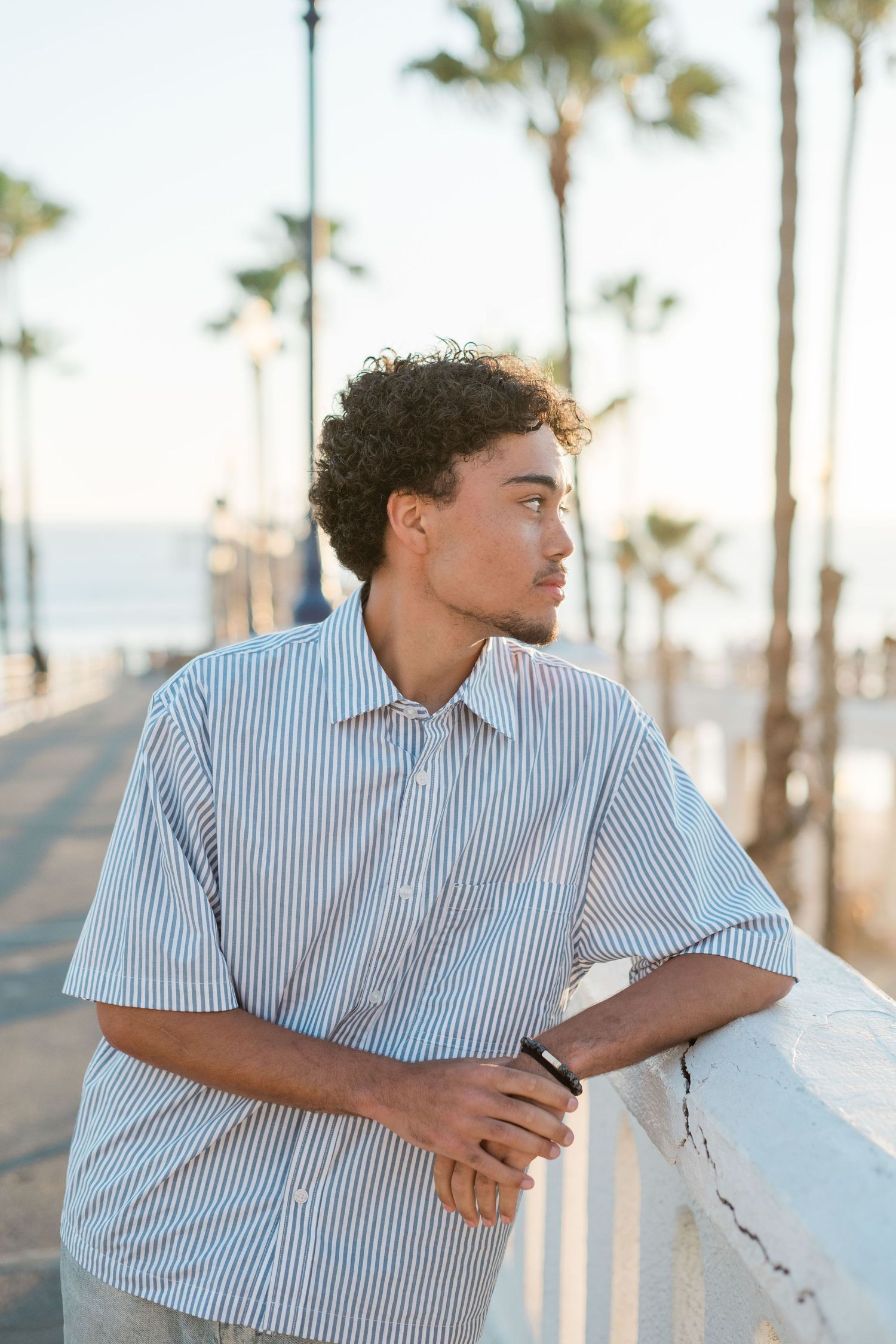 Young man in striped shirt leaning on railing at beachside promenade with palm trees at sunset.
