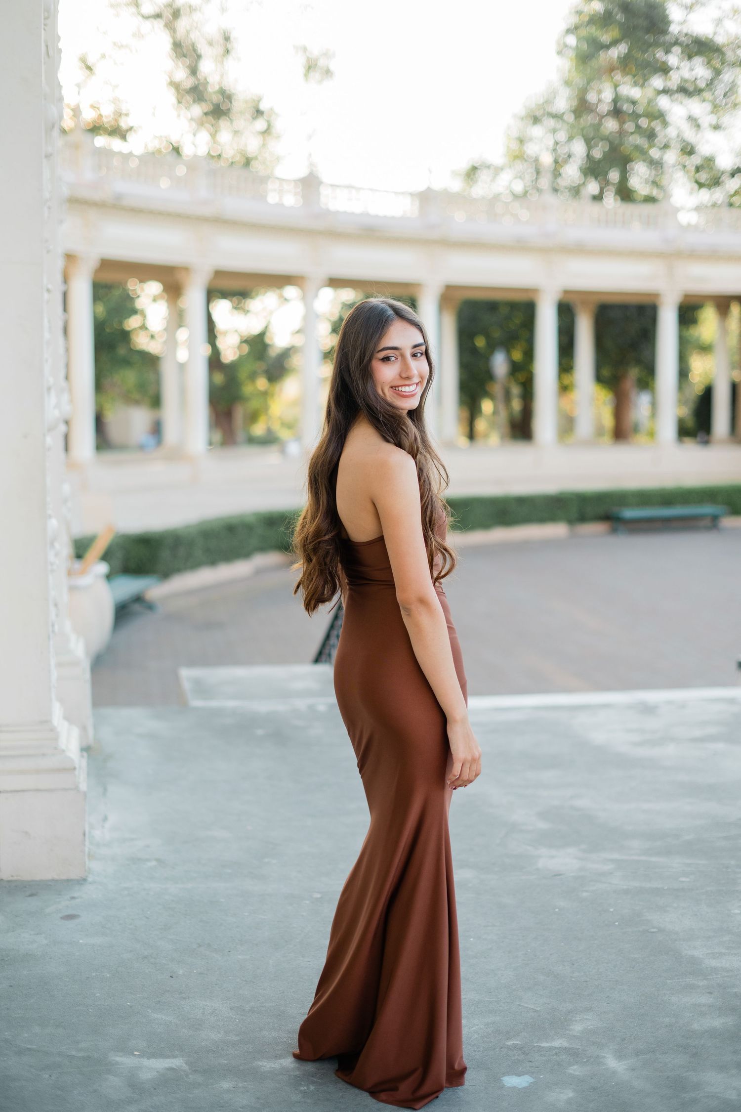 Young woman in elegant brown maxi dress smiling outdoors near classical white columns in a park setting.