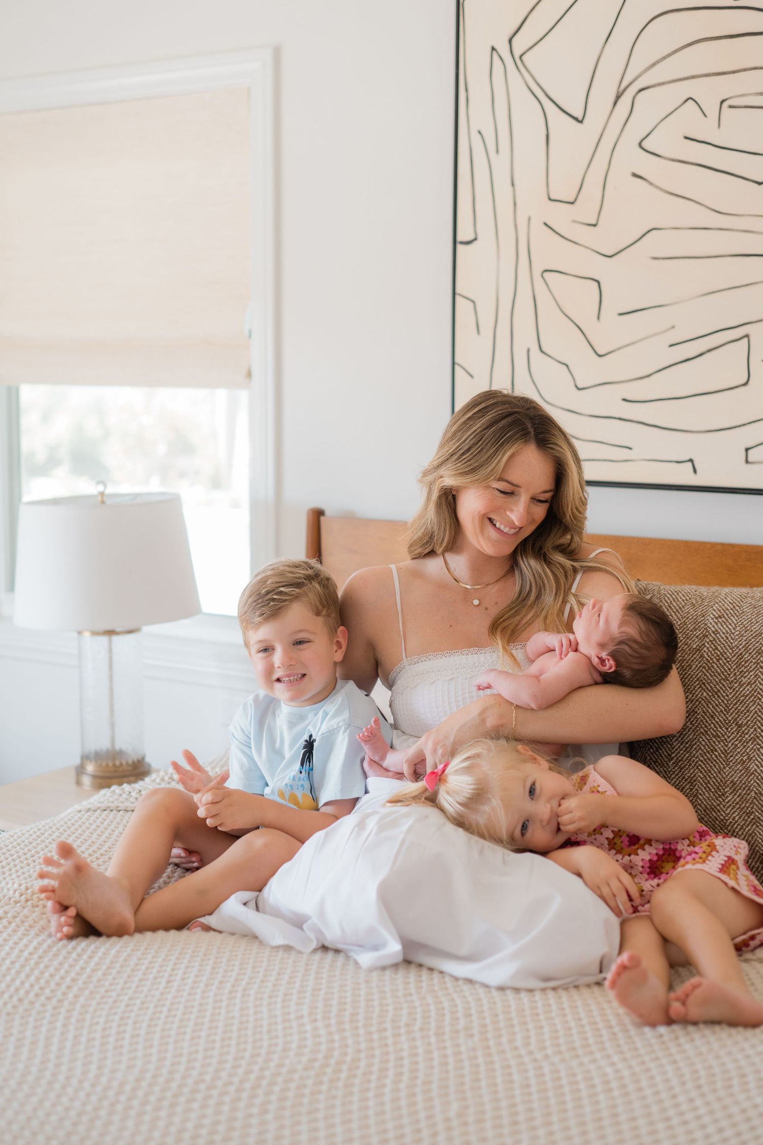 A smiling mother sits on a bed holding a newborn while three young children cuddle around her in a bright bedroom.