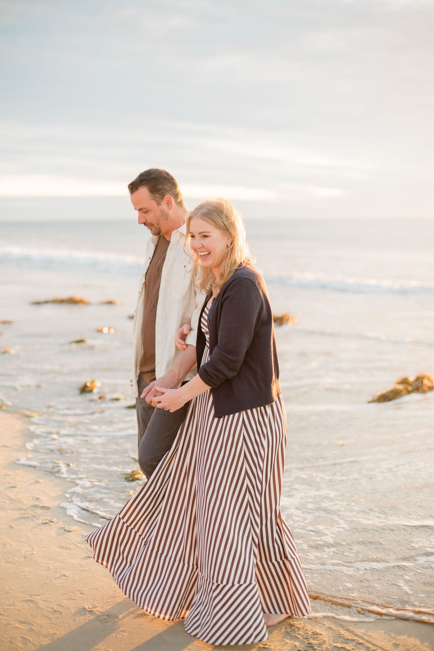 A smiling blonde woman in a striped maxi skirt walks hand-in-hand with a man along a sunny beach.